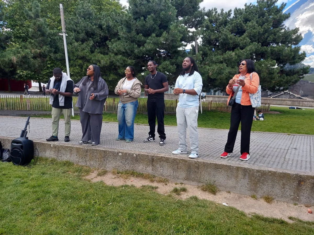 Singers at an outdoor community event beside trees and a portable speaker.