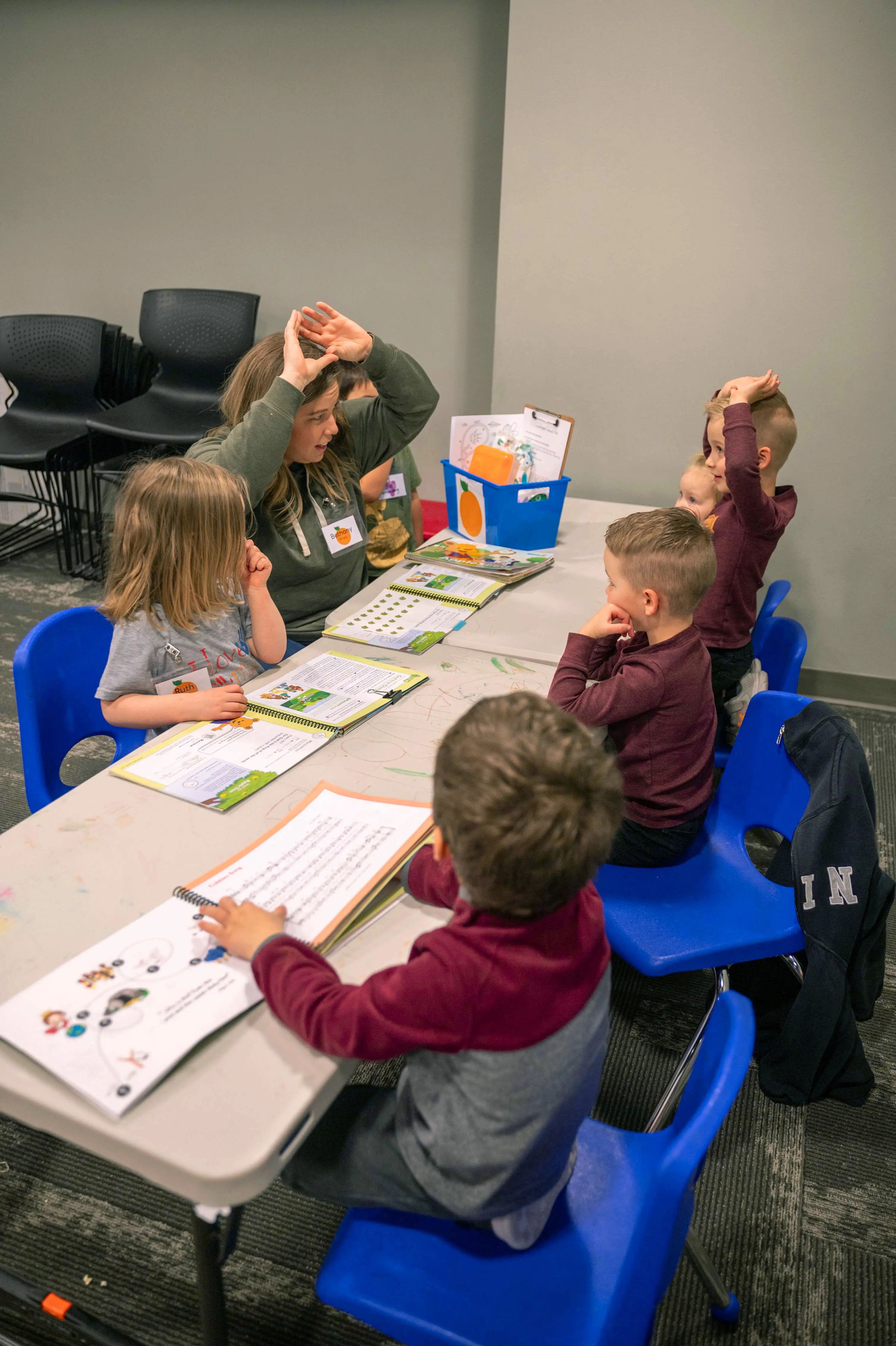 A female volunteer leads a group of young children in a lesson at Walnut Creek Kids Ministry. The group is seated at a long white table with open workbooks. The volunteer and one of the young boys have their hands raised to their heads, mimicking a roof or mountain shape as part of an interactive activity. The children are seated in bright blue plastic chairs in a modern classroom with grey walls and carpeted floors. A blue bin with an orange logo and various teaching supplies sits on the table.