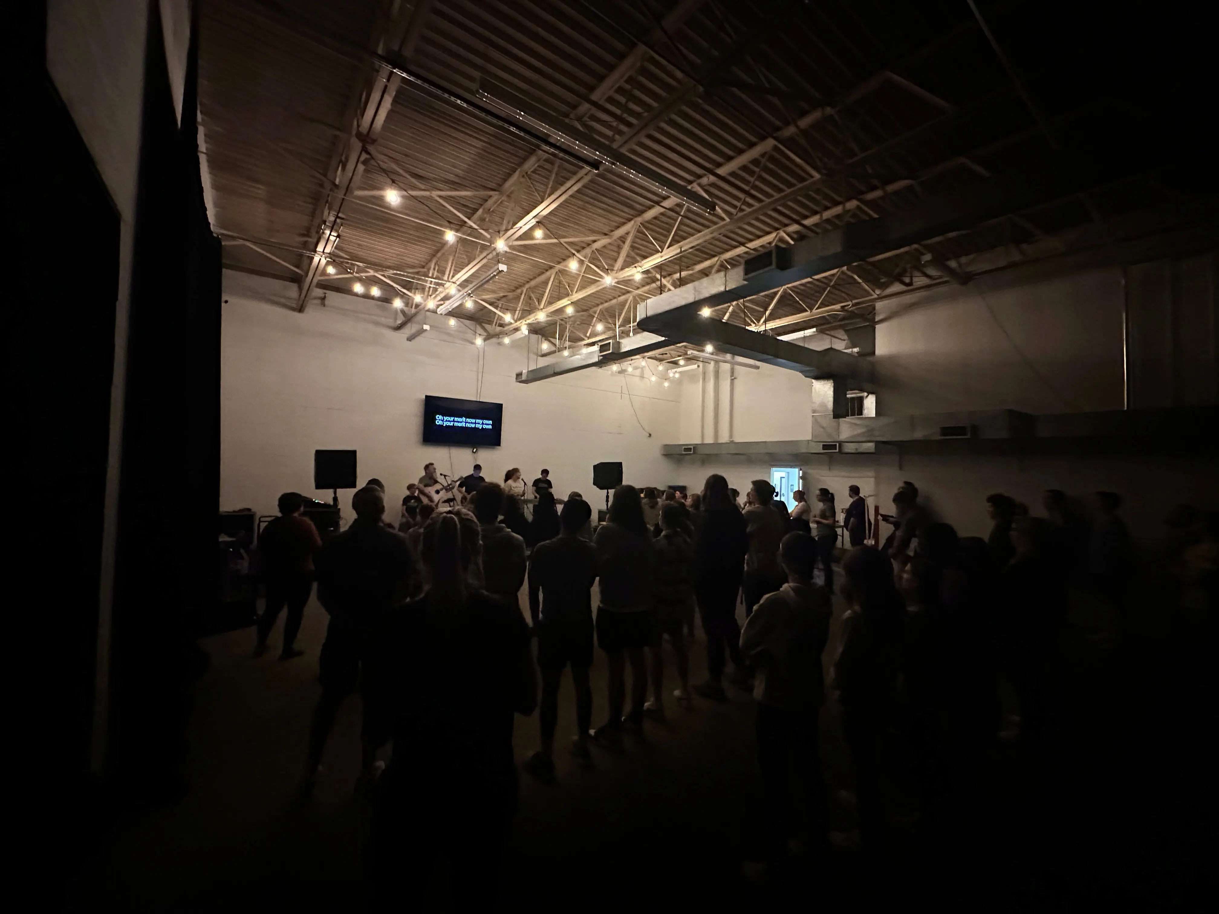 A large group of young people gathered for a worship service in an industrial-style room at Walnut Creek Downtown. The group is silhouetted against a bright white wall where a band is performing on a small stage. A TV screen displays song lyrics, and warm string lights hang from the exposed metal rafters of the high ceiling. The atmosphere is intimate and focused, with the participants standing together in the darkened space.