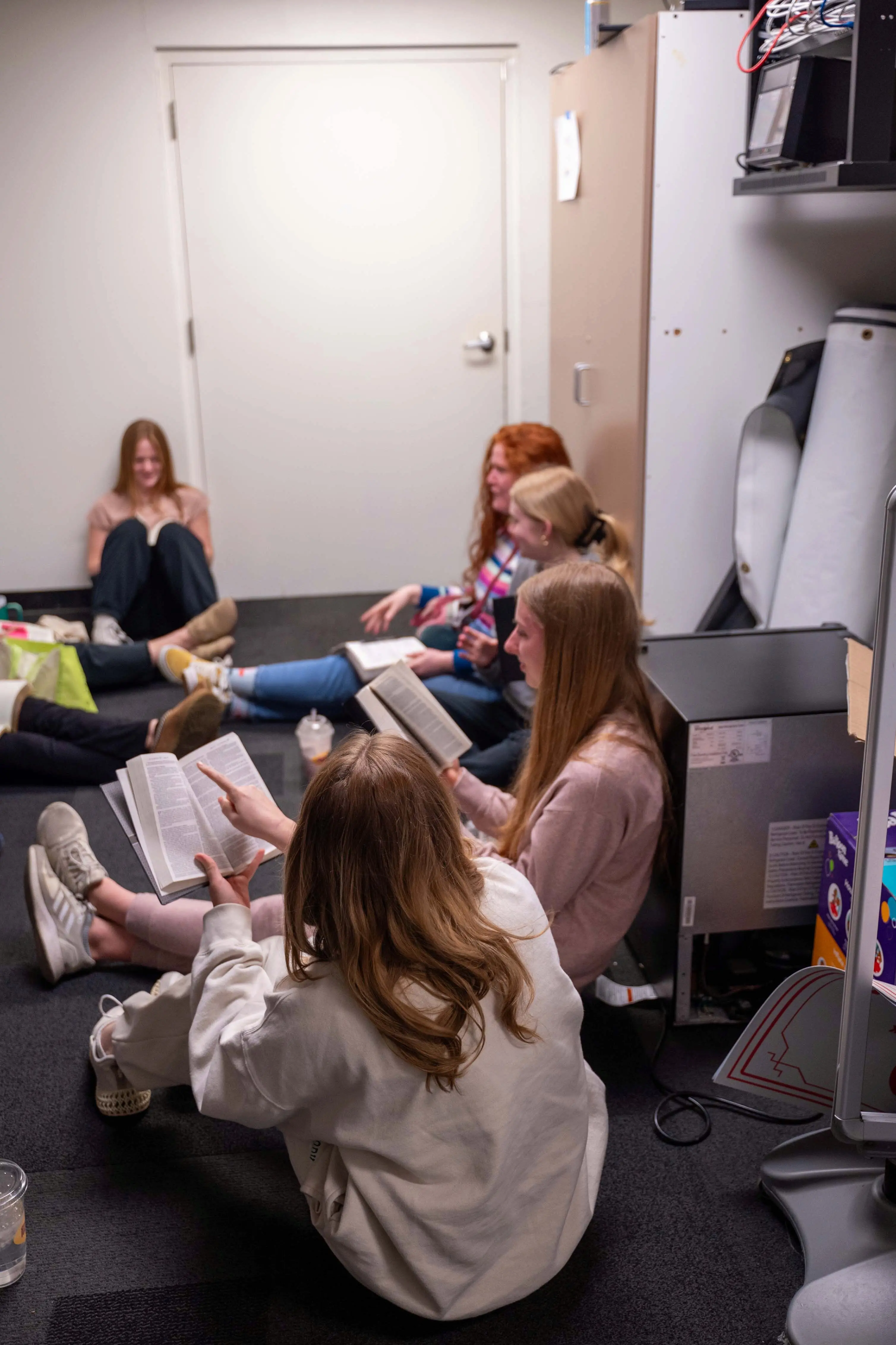 A small group of young women sit on the floor of a brightly lit room for a Bible study at Walnut Creek Downtown. The girls are gathered in a circle, leaning against the walls and furniture, each with an open Bible in their lap. One girl in the foreground, wearing a white sweatshirt, points to a passage in her Bible as she shares with the group. The room appears to be a casual meeting or storage space, featuring neutral-toned walls, dark carpeting, and a tall wooden cabinet in the background.