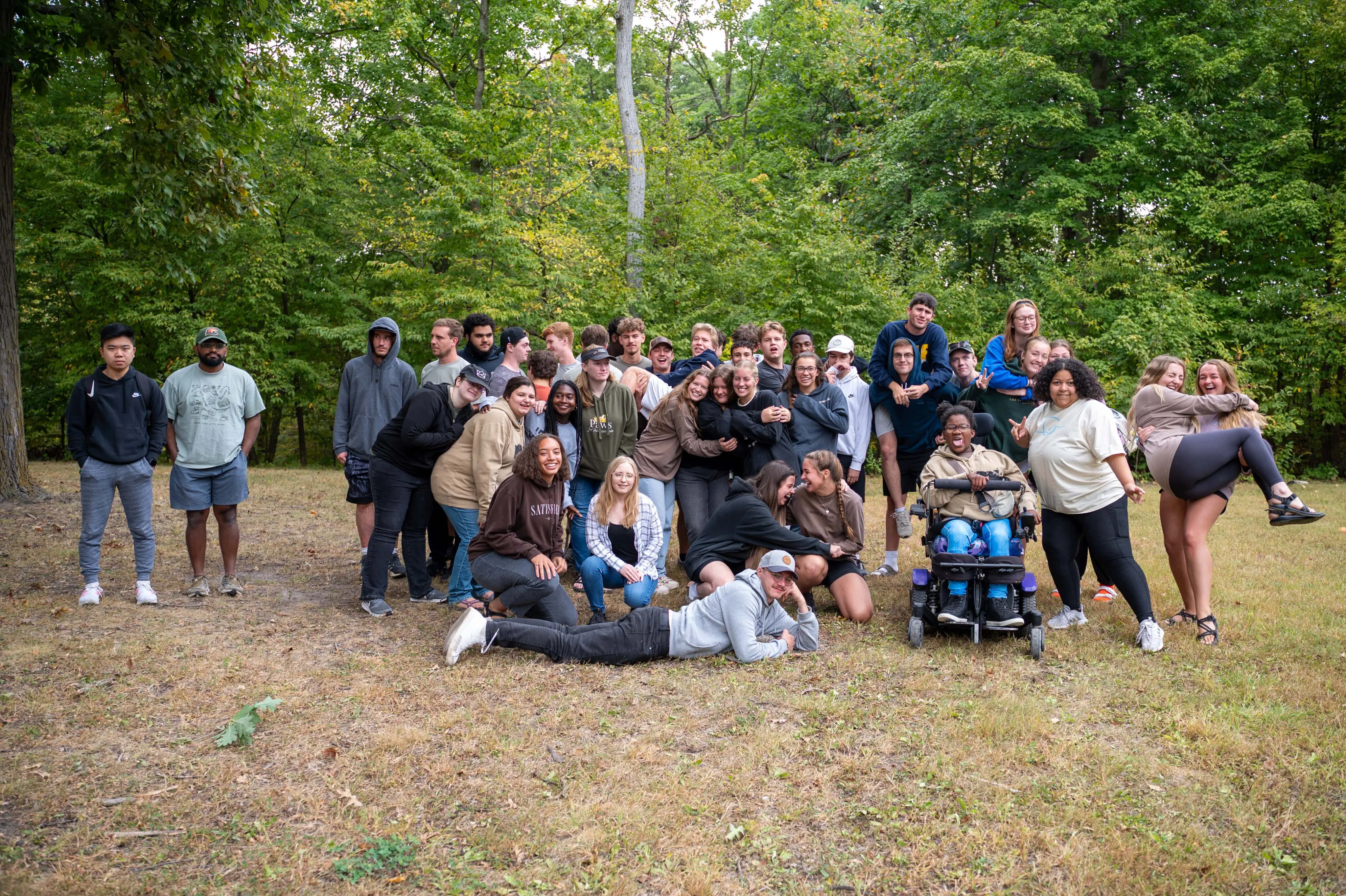 A large, diverse group of young adults poses for a playful group photo in a grassy clearing with a dense forest of green trees in the background. The group is gathered closely together, with some people hugging, one person in a motorized wheelchair smiling in the center, and a man lying casually on the grass in the foreground. To the left, two men stand slightly apart from the main group. The atmosphere is casual and joyful, with many individuals wearing hoodies and outdoor attire.