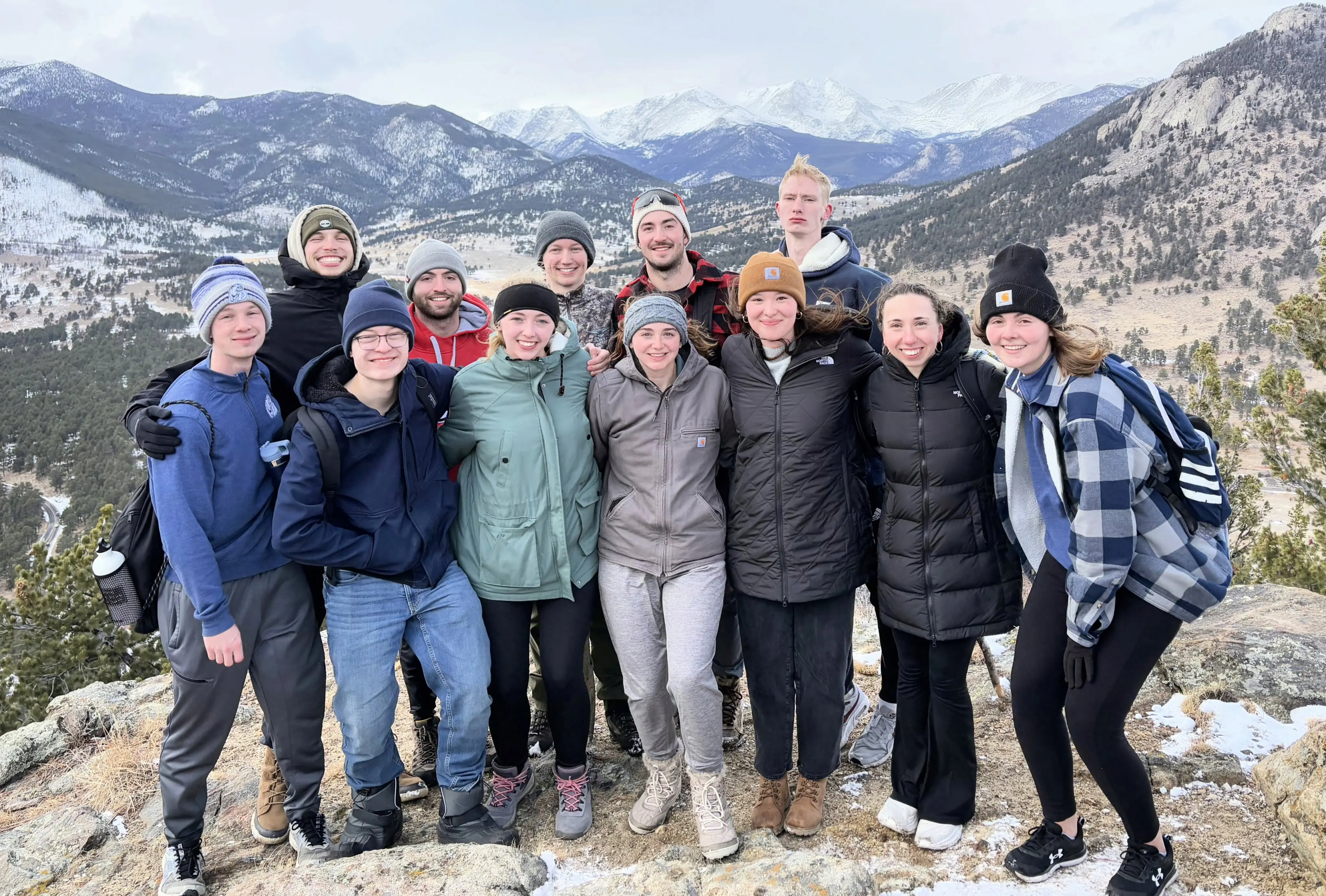 A group of twelve young men and women stand close together on a rocky, snow-dusted overlook for a group photo. They are all dressed in winter gear, including parkas, beanies, and hiking boots. Behind them, a vast mountain landscape stretches out under an overcast sky, featuring snow-capped peaks and evergreen-covered valleys. The group is smiling and huddled together against the cold, capturing a scenic moment during a group outing.