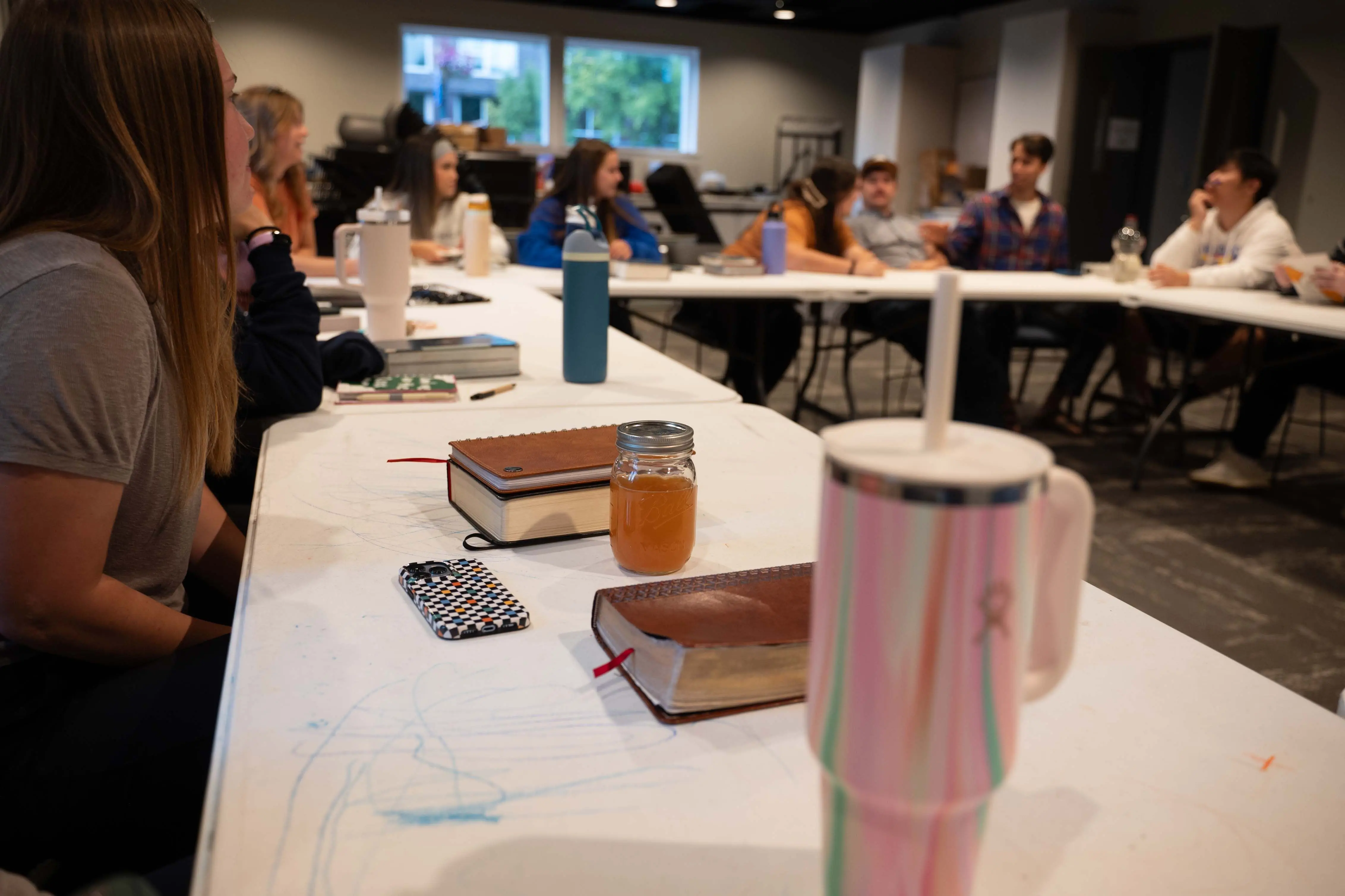 A group of young adults sit around several long white tables arranged in a "U" shape for a Bible study. The foreground features a close-up of a table with leather-bound Bibles, a mason jar with a drink, and a colorful insulated tumbler. In the blurred background, the participants are engaged in conversation, some looking toward a young man who is speaking. The room is spacious with neutral walls and a window in the distance.