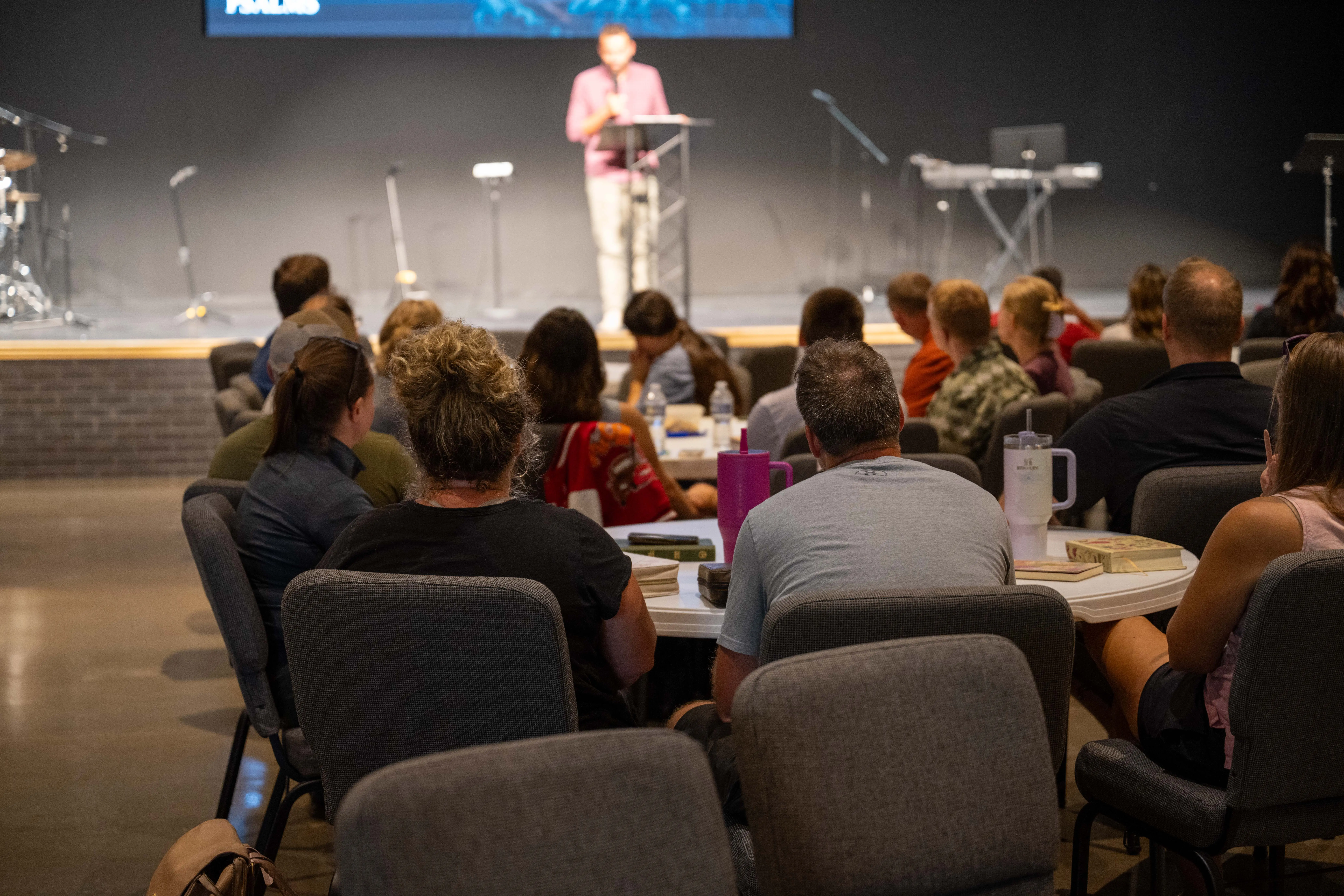 A large group of people gathers for a Wednesday night Bible study at Walnut Creek Downtown. The attendees are seated at round tables and in rows of chairs, facing a stage where a man stands at a lectern speaking. The room has a modern, industrial feel with dark walls and a large screen displaying the word "Psalms." Musical instruments, including a drum kit and keyboard, are visible on the stage behind the speaker, and several Bibles and water bottles are placed on the tables in the foreground.