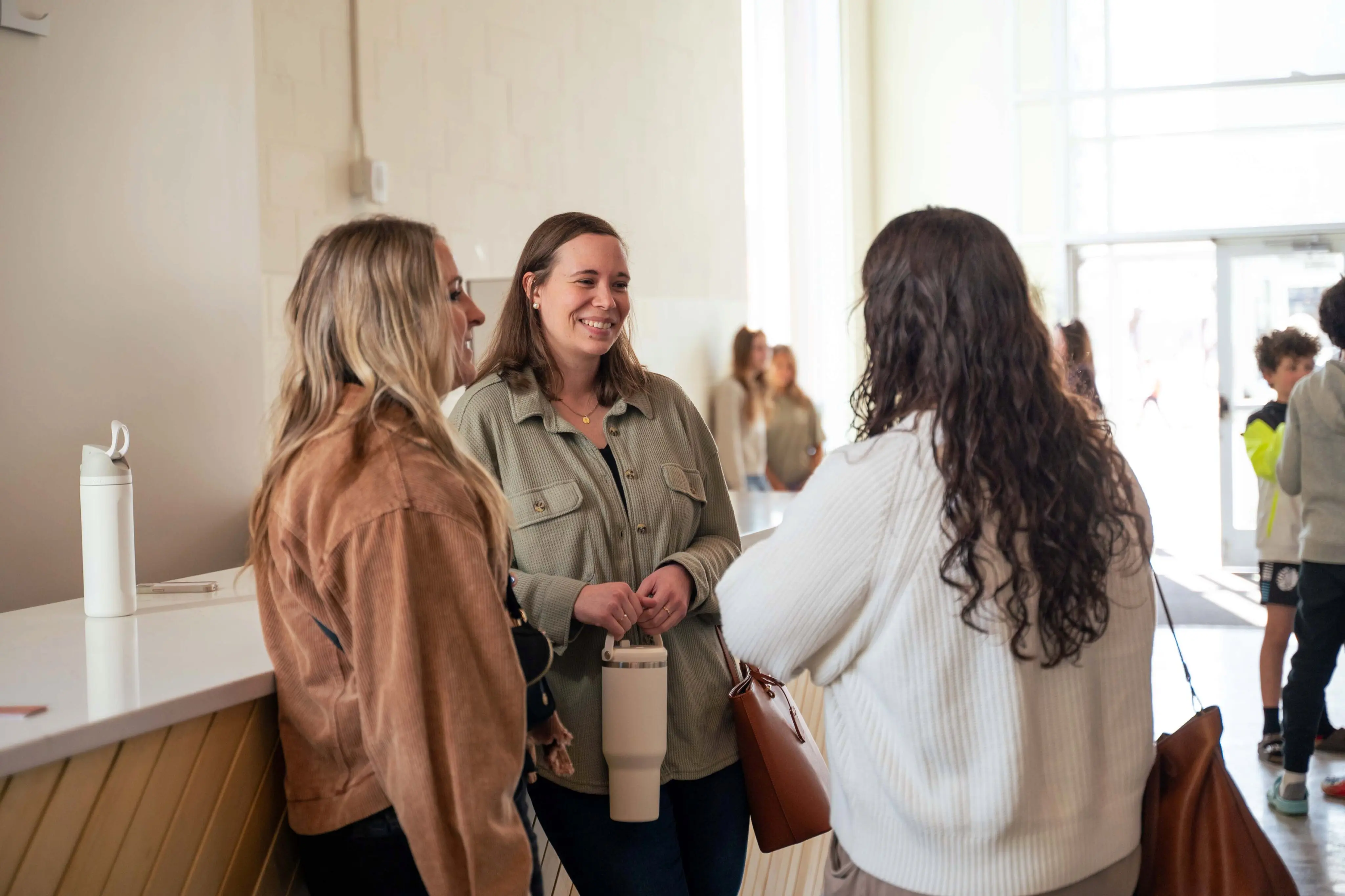 A group of young adults connecting and smiling at the Welcome Desk in the modern, light-filled atrium of Walnut Creek Church Downtown. The image shows three women talking casually near a white countertop, featuring a reusable water bottle and a handbag, with other community members and a sunlit entrance in the background, highlighting the church's welcoming atmosphere and communal spaces.
