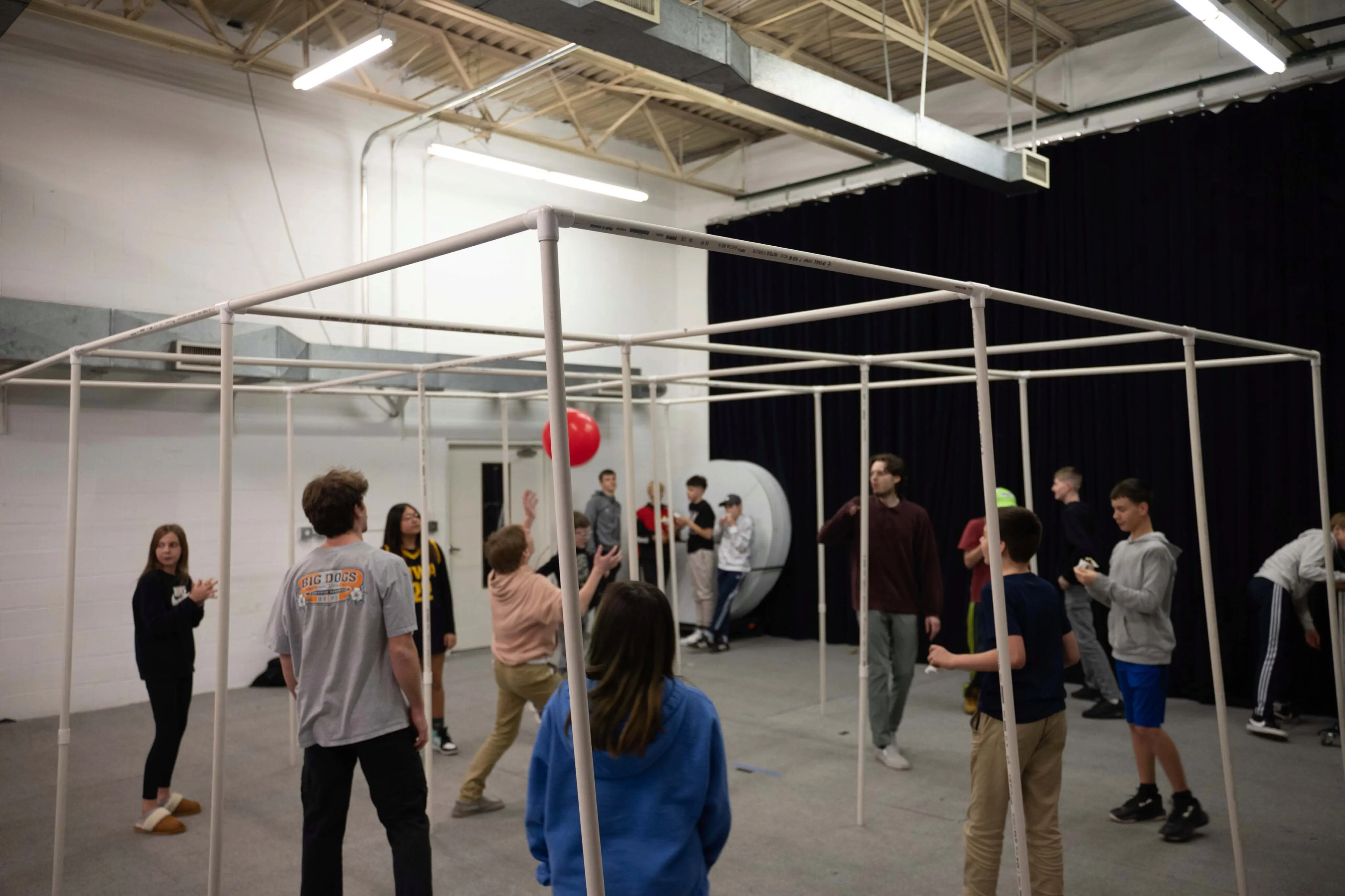 A group of teenagers plays a game of "9 Square in the Air" inside a gymnasium at Walnut Creek Downtown. The game is played within a large, square grid structure made of white PVC piping. A red ball is tossed high into the air as participants look up, ready to hit it. Several other teens stand around the perimeter of the grid, waiting for their turn to play. The room features white walls, industrial lighting, and a large black curtain in the background.