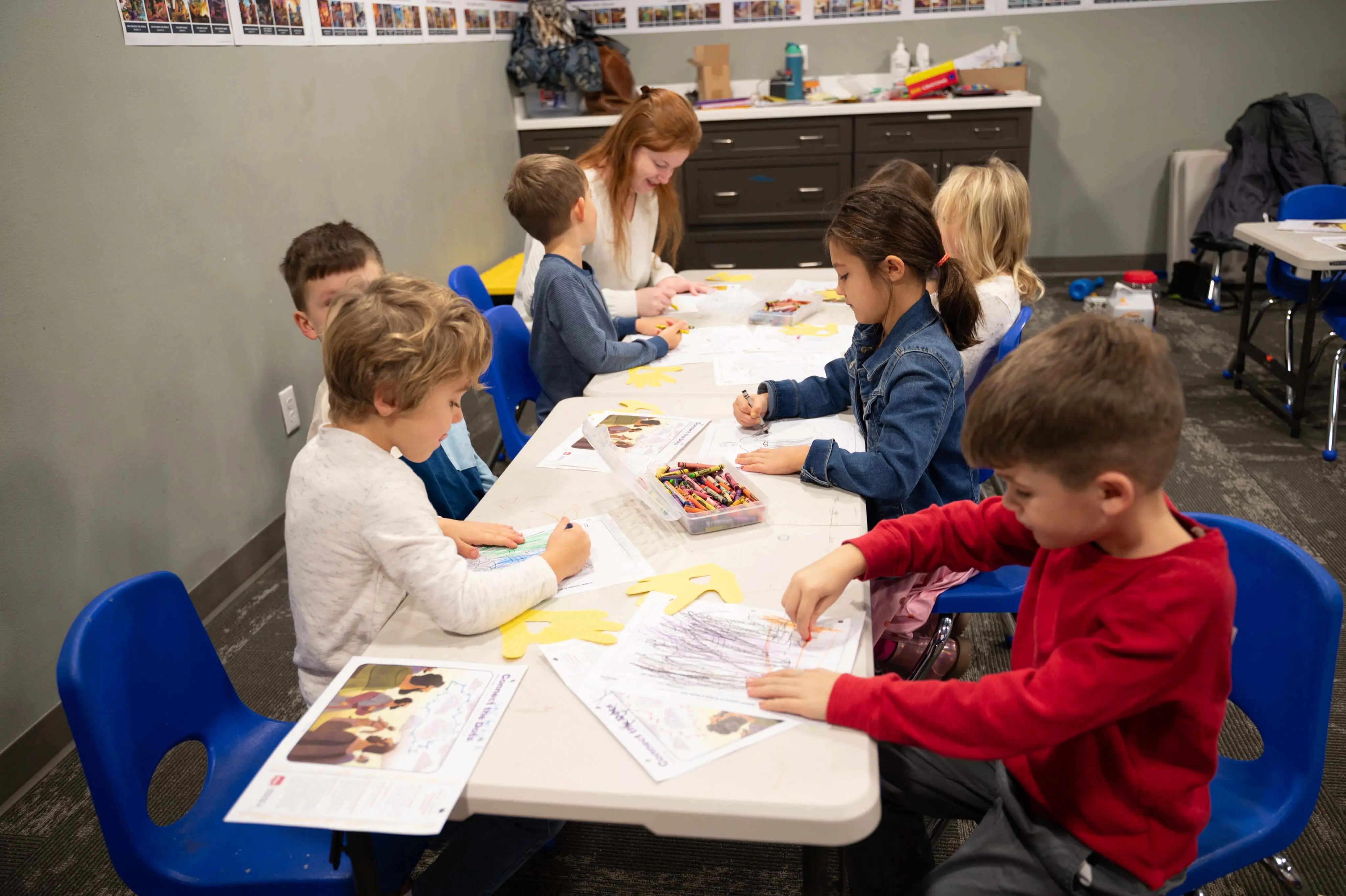 A group of young children sitting at small white tables in a classroom at Walnut Creek Downtown, coloring and working on crafts. A red-haired female volunteer sits at the back table, assisting a young boy. The children are focused on their coloring pages and yellow hand-shaped paper cutouts, with containers of crayons scattered across the tables. The room is bright with grey walls, blue plastic chairs, and a row of colorful pictures displayed along the top of the wall.