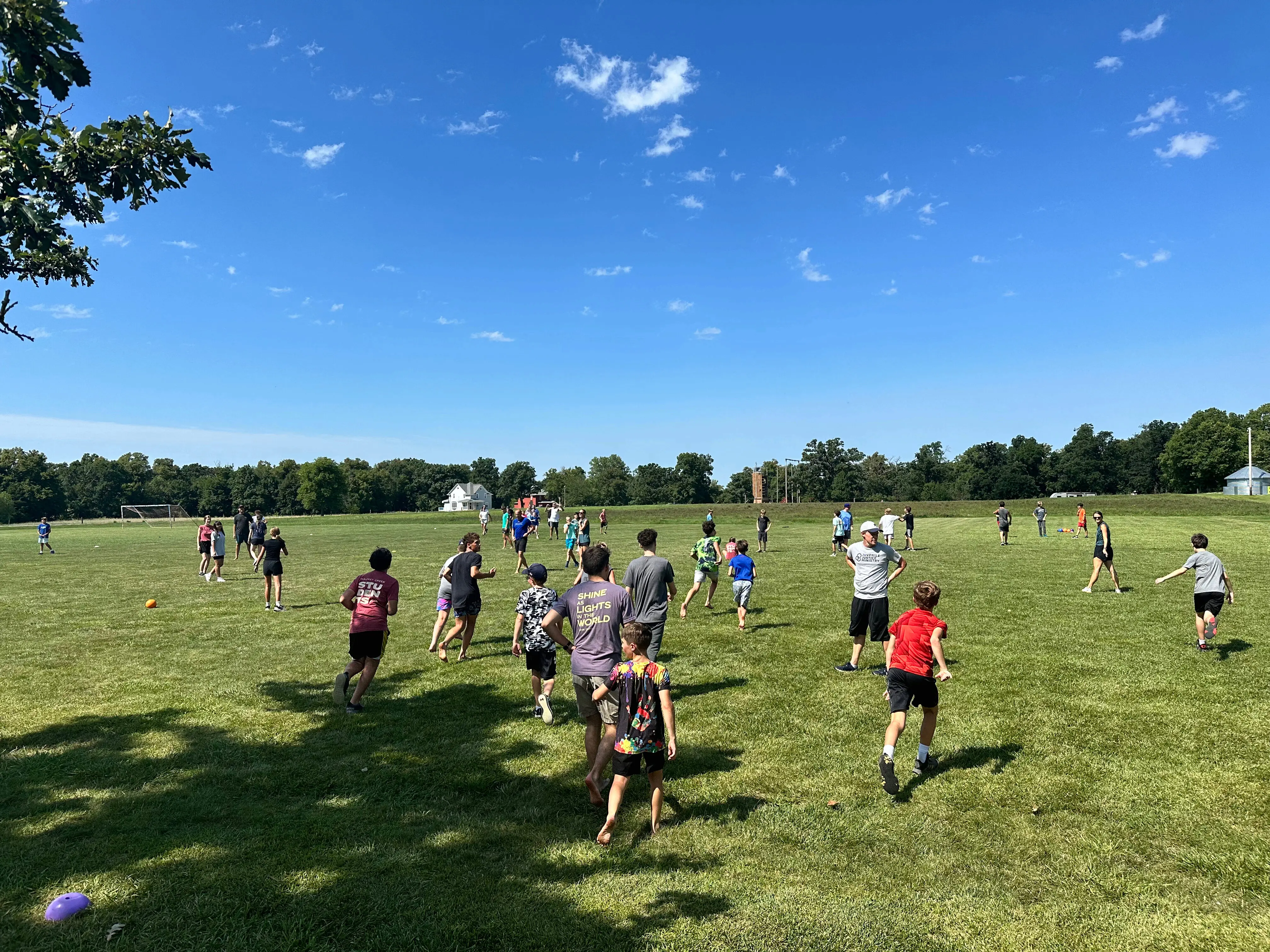 A wide-angle shot of a large group of teenagers playing an organized game on a vast green field during a sunny day at a Walnut Creek youth retreat. The participants are scattered across the grass, some running and others standing in groups, under a clear blue sky with light, wispy clouds. A line of trees and a small white house are visible in the far distance. In the foreground, several teens are seen from behind, including one wearing a purple shirt that says "Shine as Lights in the World."