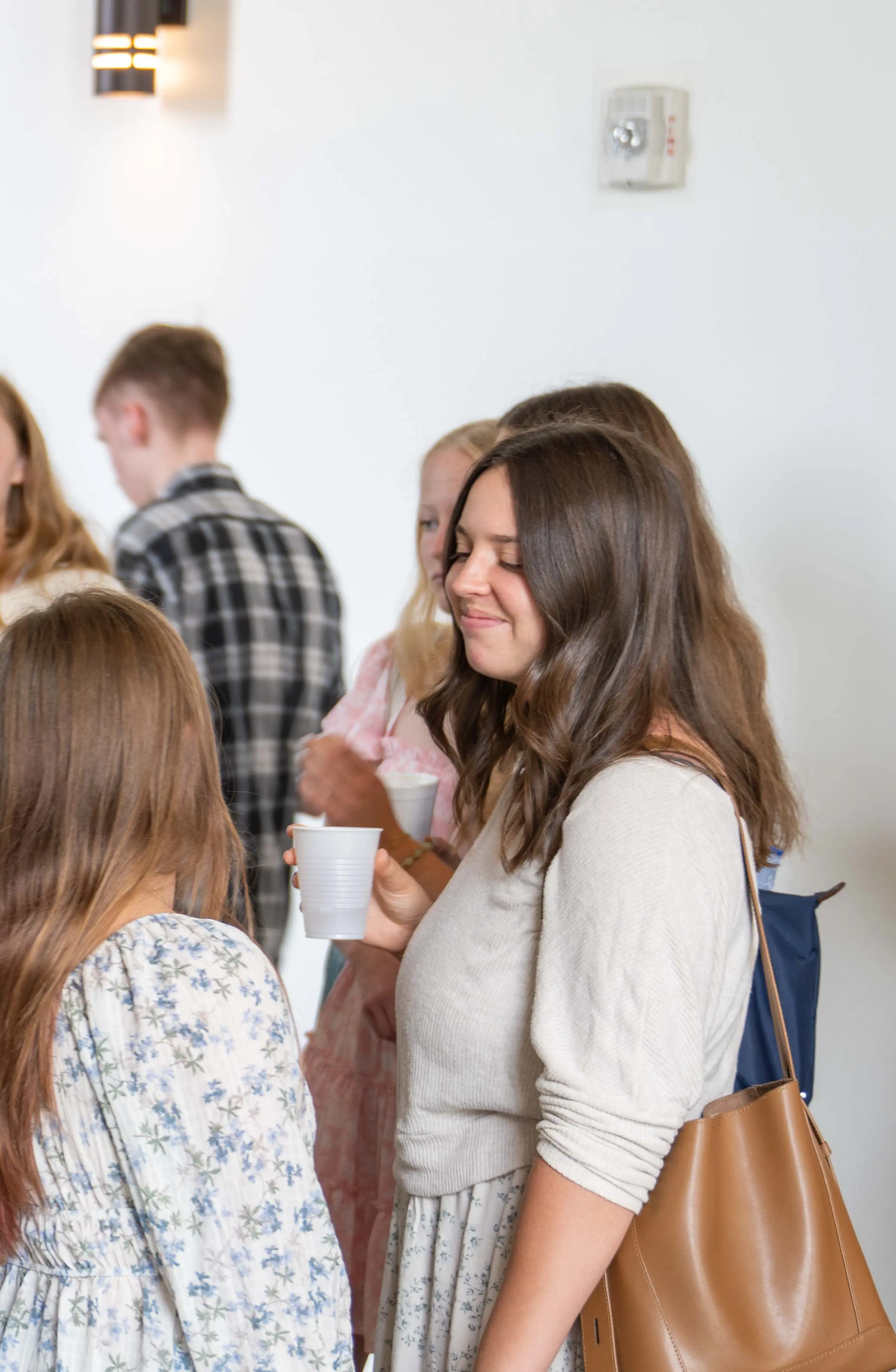 A group of young adults connecting and smiling at the Welcome Desk in the modern, light-filled atrium of Walnut Creek Church Downtown. The image shows three women talking casually near a white countertop, featuring a reusable water bottle and a handbag, with other community members and a sunlit entrance in the background, highlighting the church's welcoming atmosphere and communal spaces.