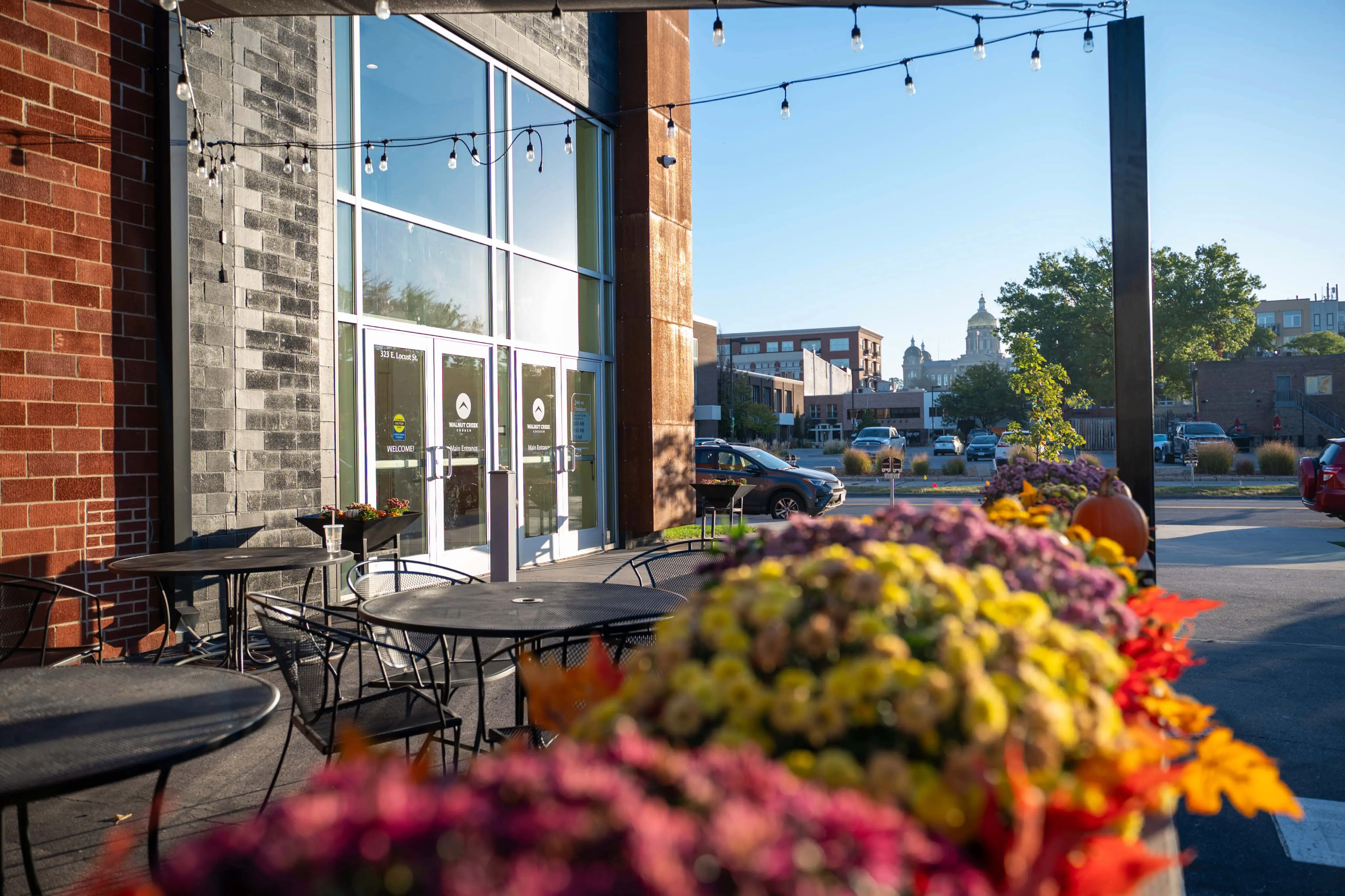 A view of the outdoor patio at Walnut Creek Downtown on a clear, sunny day. The foreground is filled with a colorful, out-of-focus arrangement of autumnal flowers and a small pumpkin. Beyond the flowers, black metal patio tables and chairs sit beneath a string of Edison lights. The church’s modern brick and glass entrance is visible, and in the far distance, the golden dome of the Iowa State Capitol rises above the downtown Des Moines skyline.