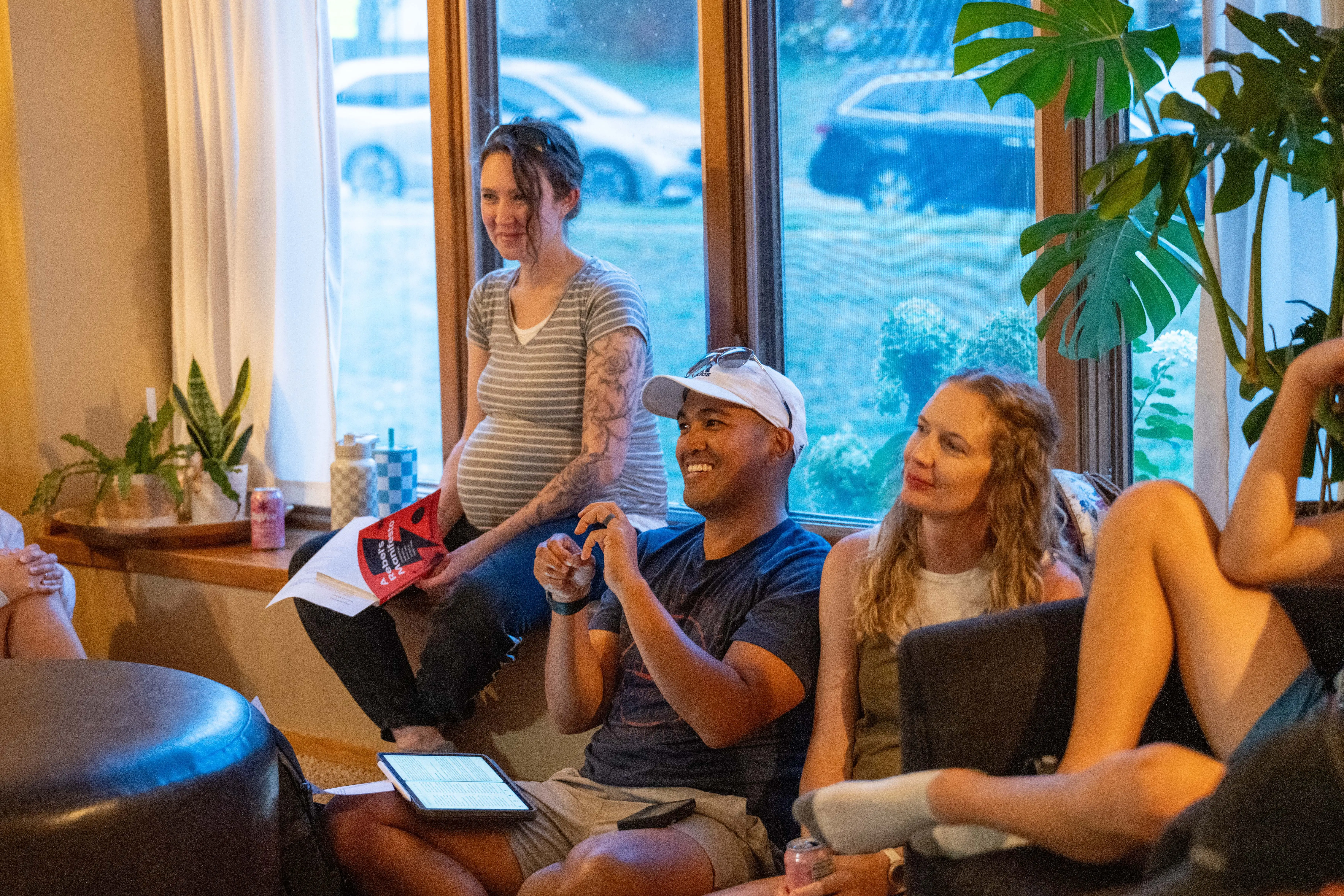 A group of adults gathered in a cozy, sunlit living room for a family-oriented home Bible study. A pregnant woman in a striped shirt sits on a window bench, smiling toward someone off-camera, while a man in a white baseball cap and a blue t-shirt sits on the floor, gesturing during a conversation. Another woman sits beside him on the floor, listening intently.
