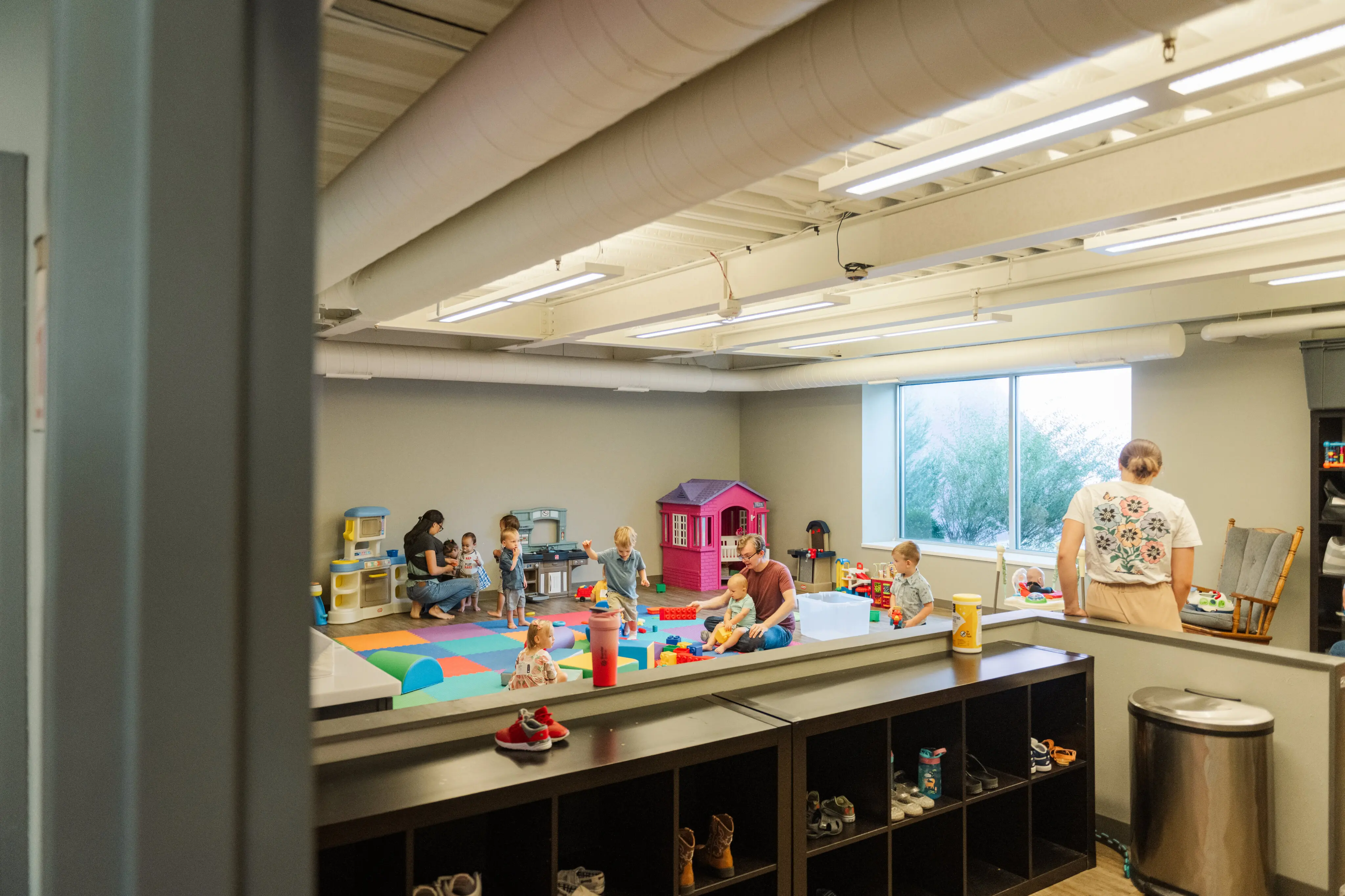 A bright and spacious nursery or kids' playroom at Walnut Creek Downtown. Several toddlers and young children are playing with colorful blocks and toys on a large foam floor mat. A few adult volunteers are interspersed among the children, including a man sitting on the floor helping a toddler and a woman standing near a cubby storage unit. The room features industrial-style ceilings, a pink toy playhouse, and a large window that lets in natural light. In the foreground, a dark wood shelving unit contains pairs of children's shoes.