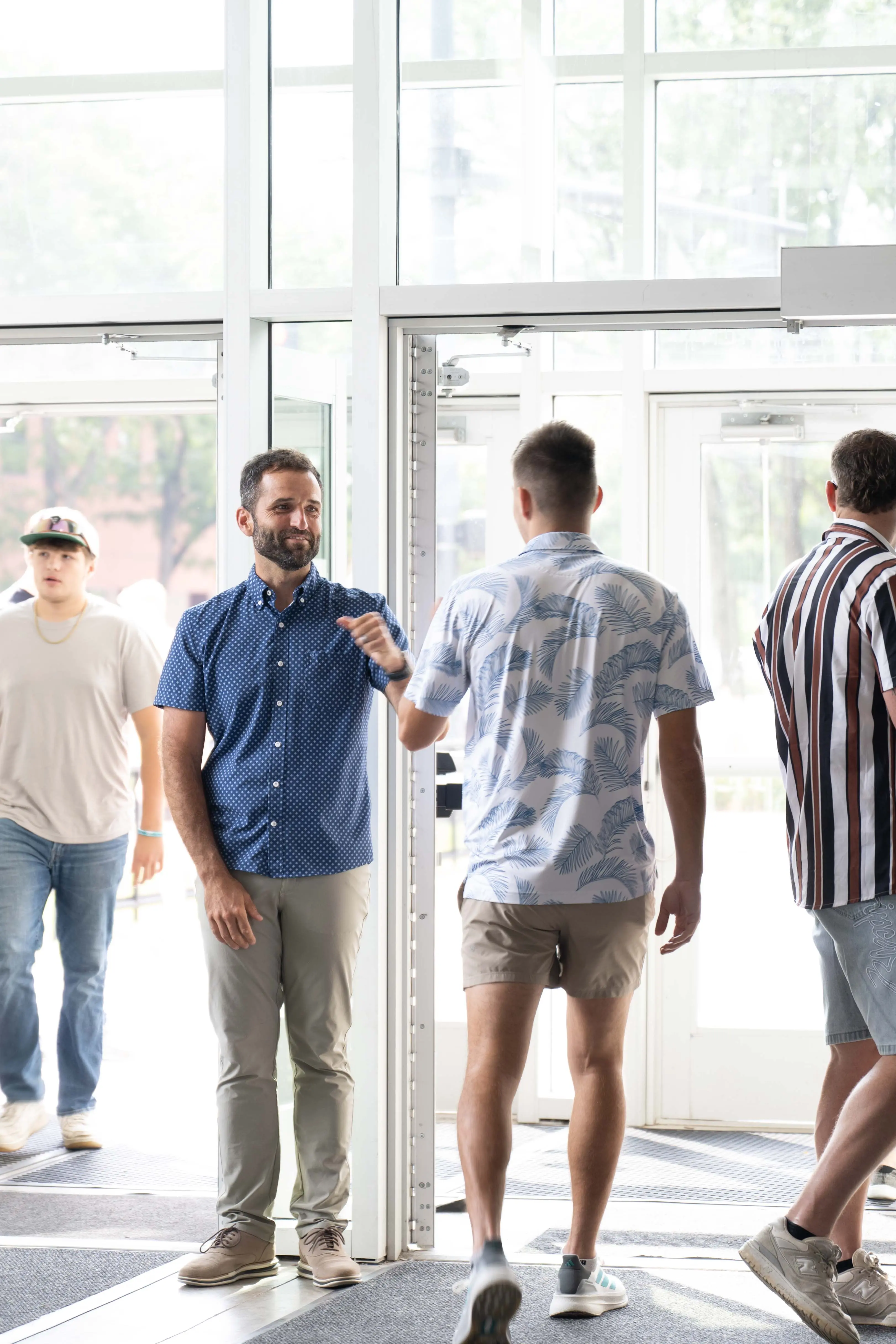 Three men talking and laughing in the bright, modern lobby of Walnut Creek Church Downtown. One man holds a toddler and a coffee tumbler while standing near the glass main entrance doors, showcasing the welcoming, family-friendly atmosphere of the church community.