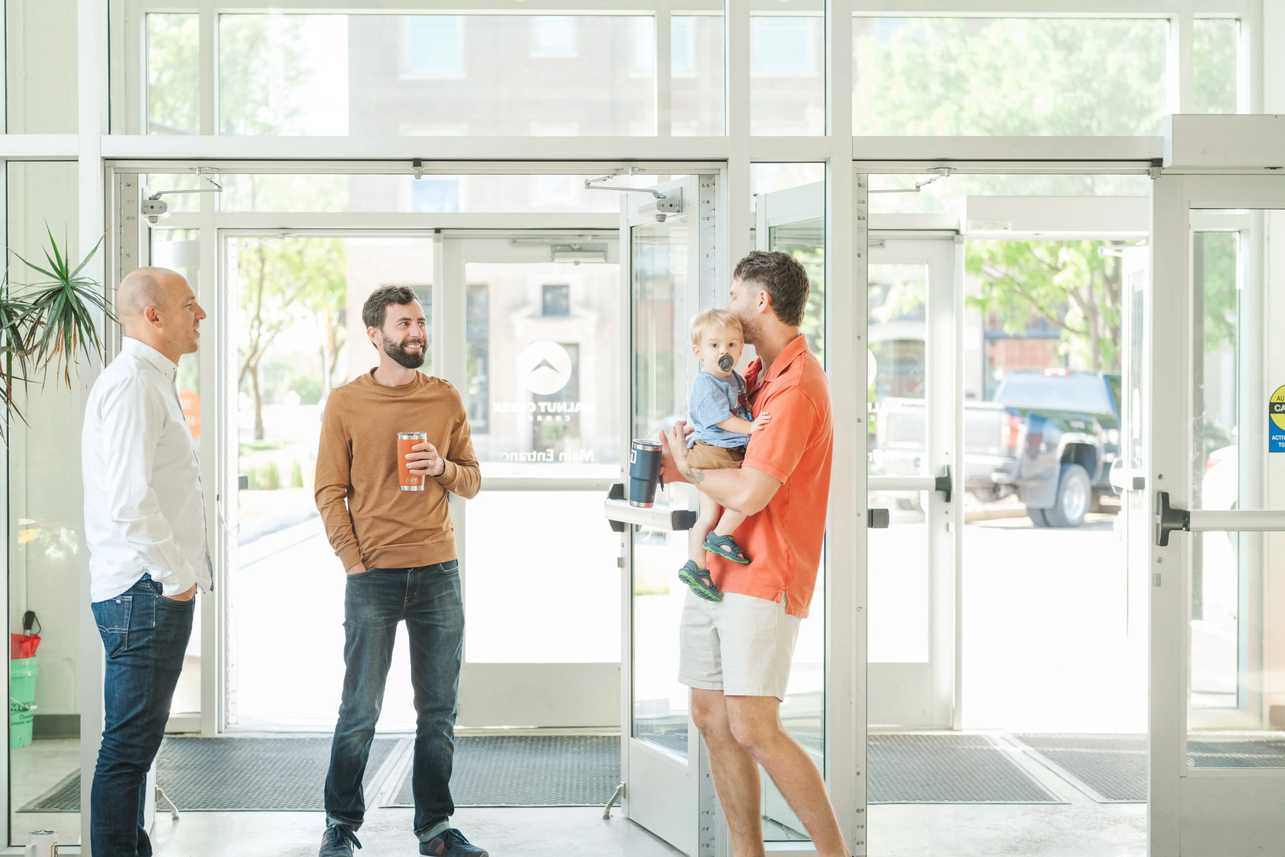 Three men talking and laughing in the bright, modern lobby of Walnut Creek Church Downtown. One man holds a toddler and a coffee tumbler while standing near the glass main entrance doors, showcasing the welcoming, family-friendly atmosphere of the church community.