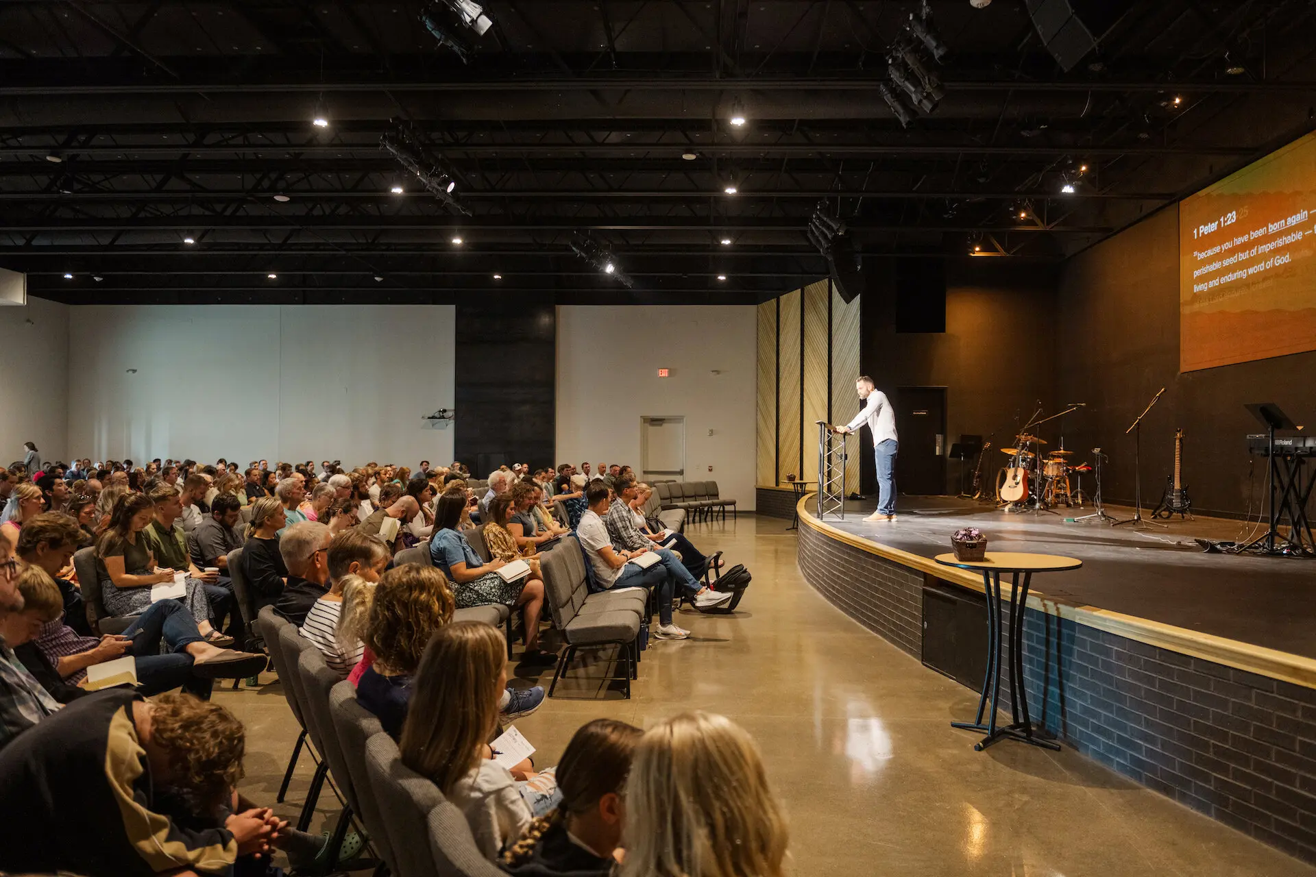 A wide-angle view of a Sunday service at Walnut Creek Downtown. A speaker stands at a podium on a raised, curved stage, addressing a large, attentive congregation seated in rows of grey chairs. To the right of the speaker, a drum set and musical equipment are visible on stage, and a large screen overhead displays the Bible verse 1 Peter 1:23-25. The auditorium has a modern, industrial feel with an exposed black ceiling, recessed lighting, and dark brick accents along the stage.