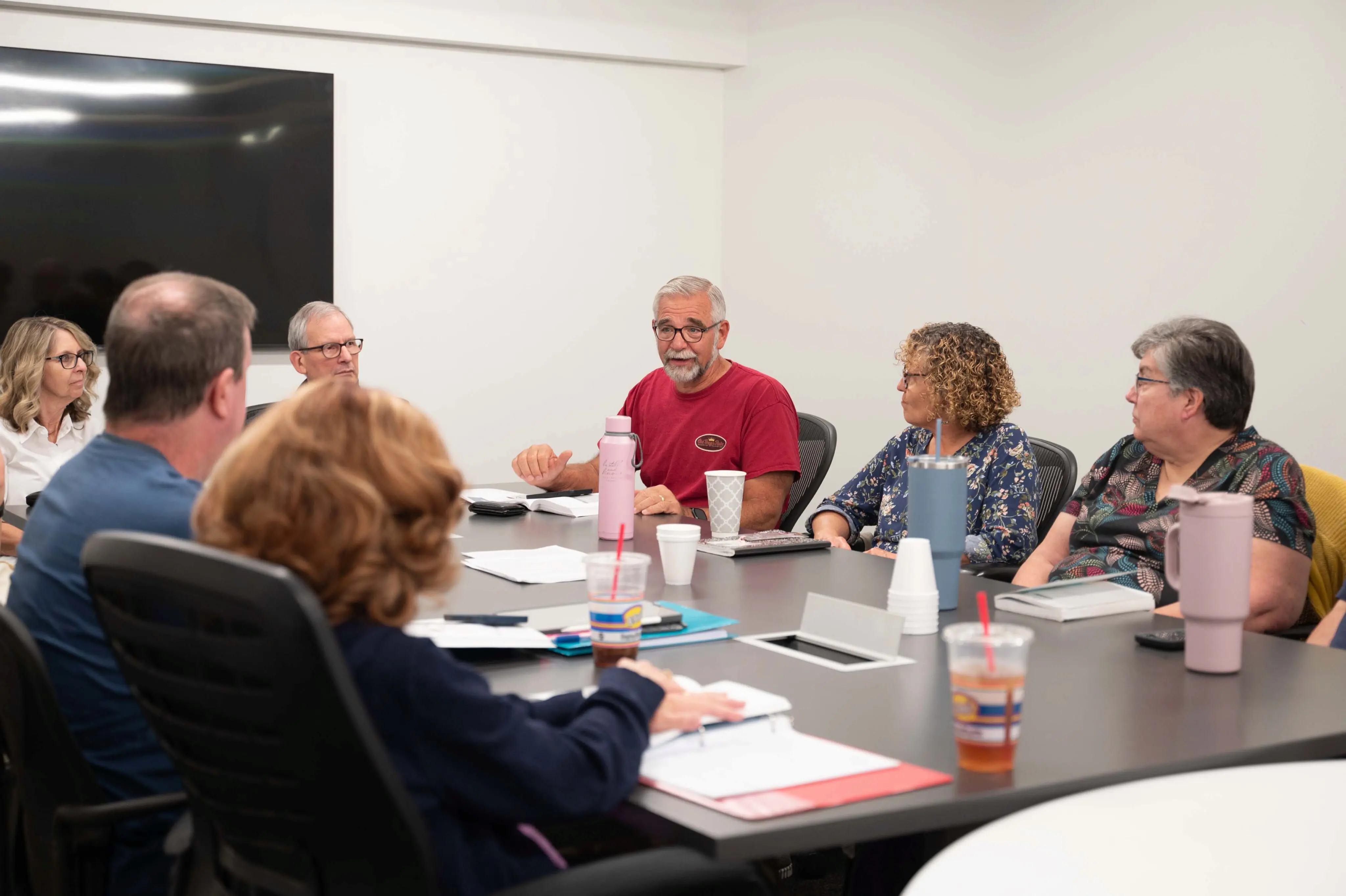 A group of adults sits around a large conference table for a Bible study. A man with grey hair and a beard, wearing a red t-shirt, speaks to the group while gesturing toward an open Bible and notebook in front of him. The other participants listen attentively, with various Bibles, notebooks, and drinks scattered across the table. The setting is a bright, modern room with white walls and a large, dark screen mounted in the background.