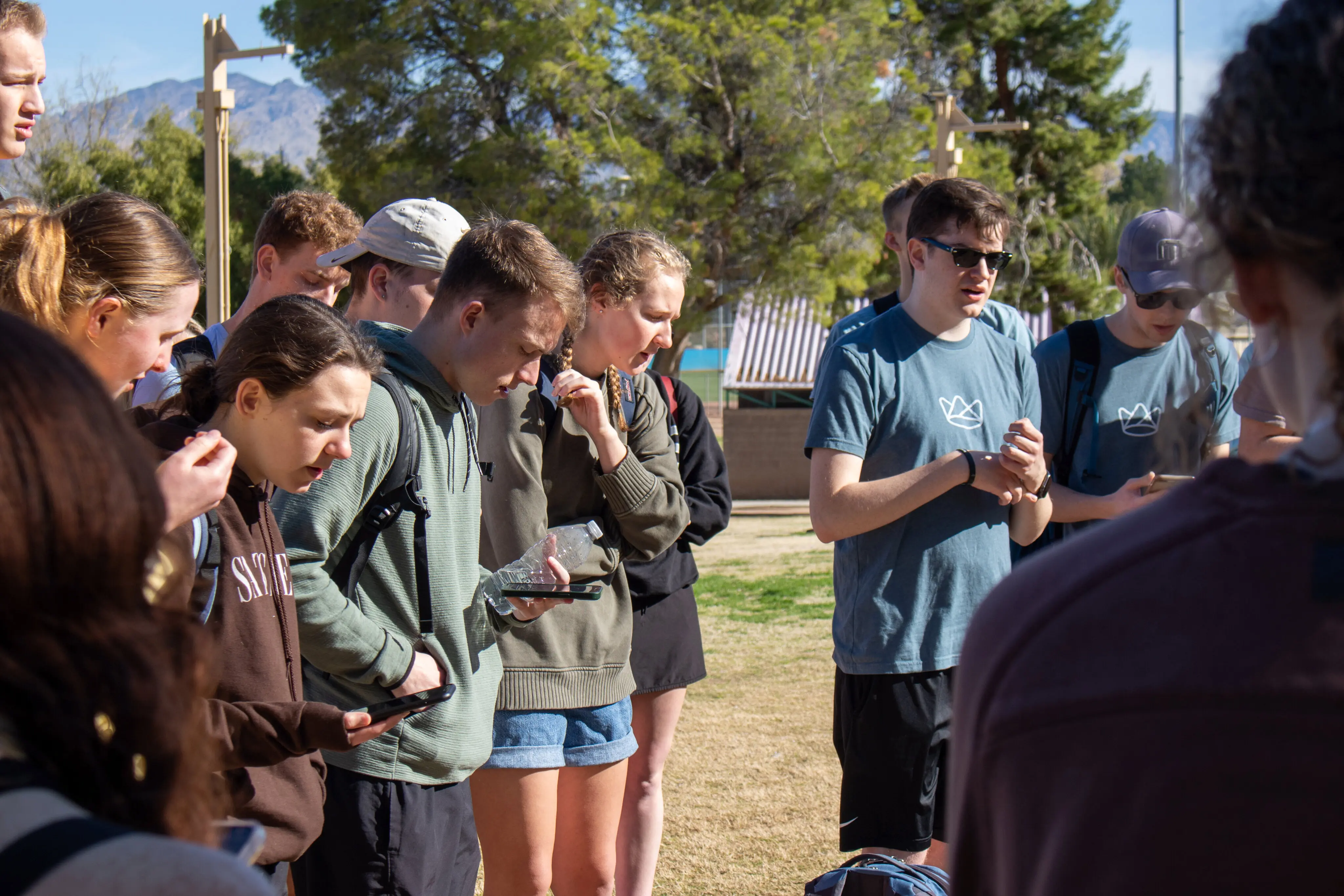 A group of young adults on a mission trip stand in a circle outdoors, bowing their heads in prayer. Some individuals hold smartphones or water bottles while others have their hands clasped. Two men in the group wear blue t-shirts featuring a small crown logo. The scene is set in a sunny, park-like area with green trees, a grassy field, and mountains visible in the distant background.