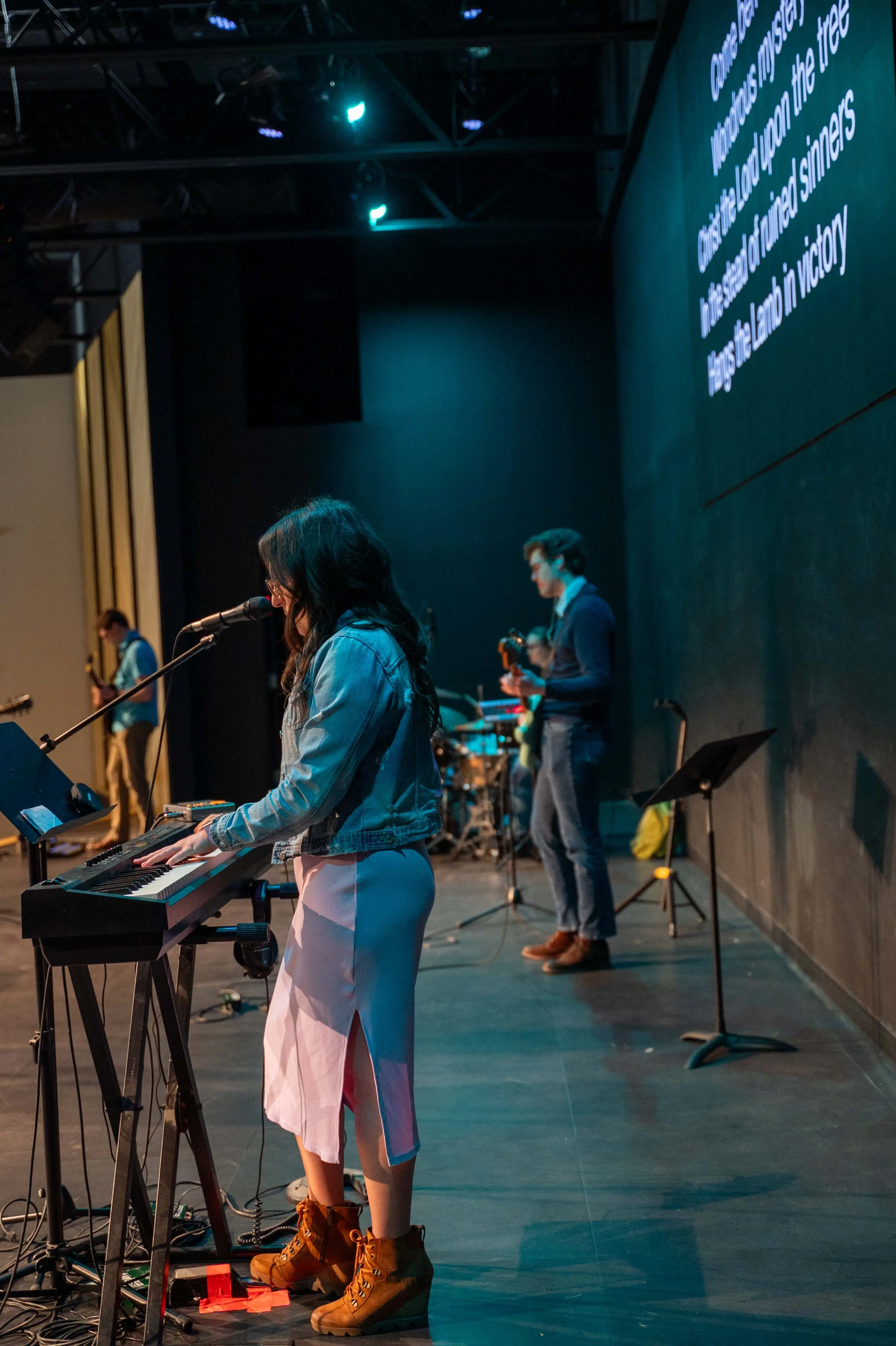 A worship band performs on stage at Walnut Creek Downtown. In the foreground, a woman with long dark hair, wearing a denim jacket, a pink dress, and brown boots, plays a keyboard and sings into a microphone. In the background, two men play guitar and bass under cool teal stage lighting. To the right, a large screen displays song lyrics starting with "Come behold the wondrous mystery." The setting is modern with dark walls and an industrial ceiling.