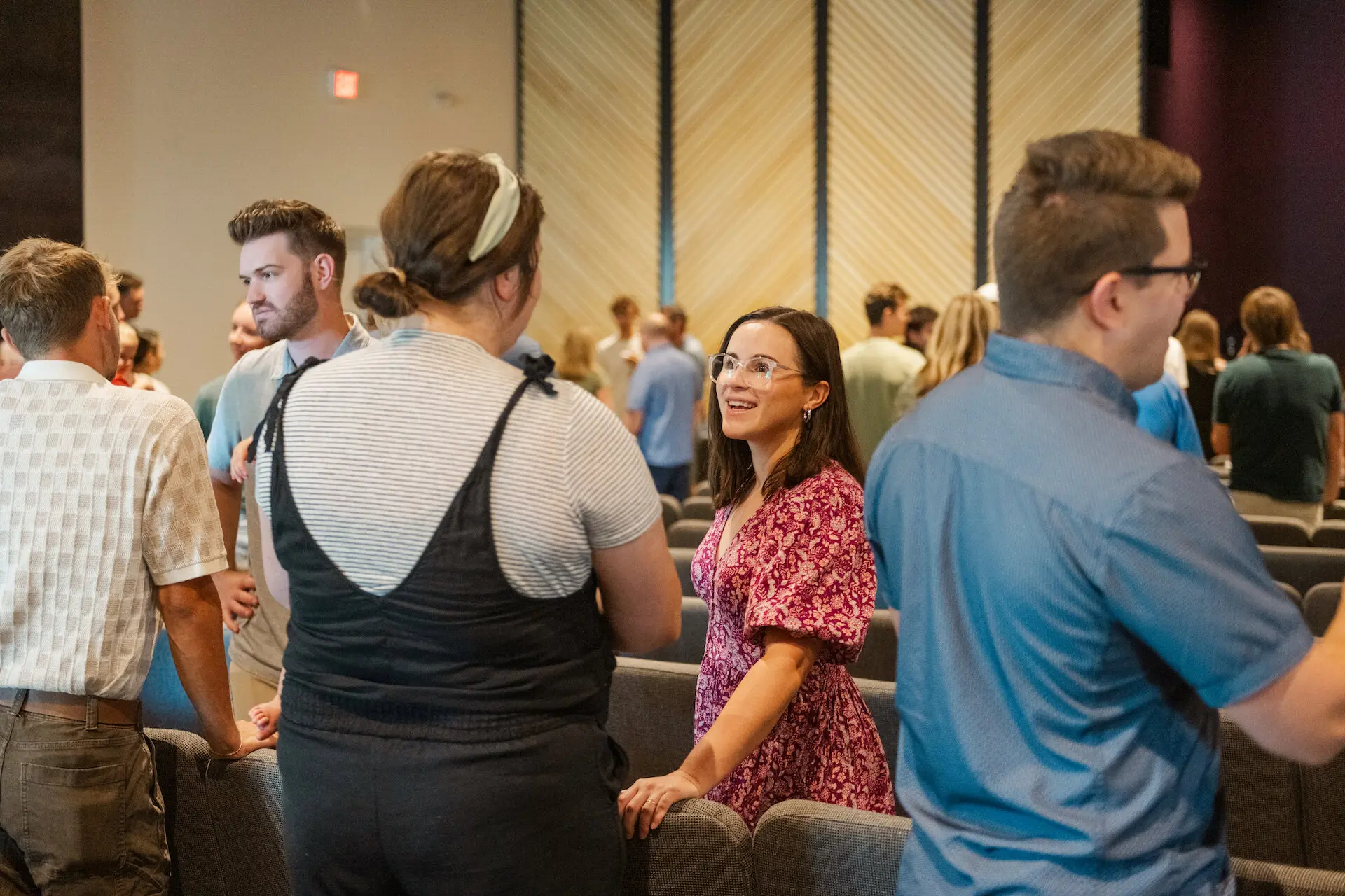 A group of people from Walnut Creek Church Downtown, including adults and children, talk warmly together in the church sanctuary. A woman in a floral dress and clear-frame glasses smiles as she speaks to another woman in overalls, highlighting the personal connections made during community events and Sunday services in a modern worship space.