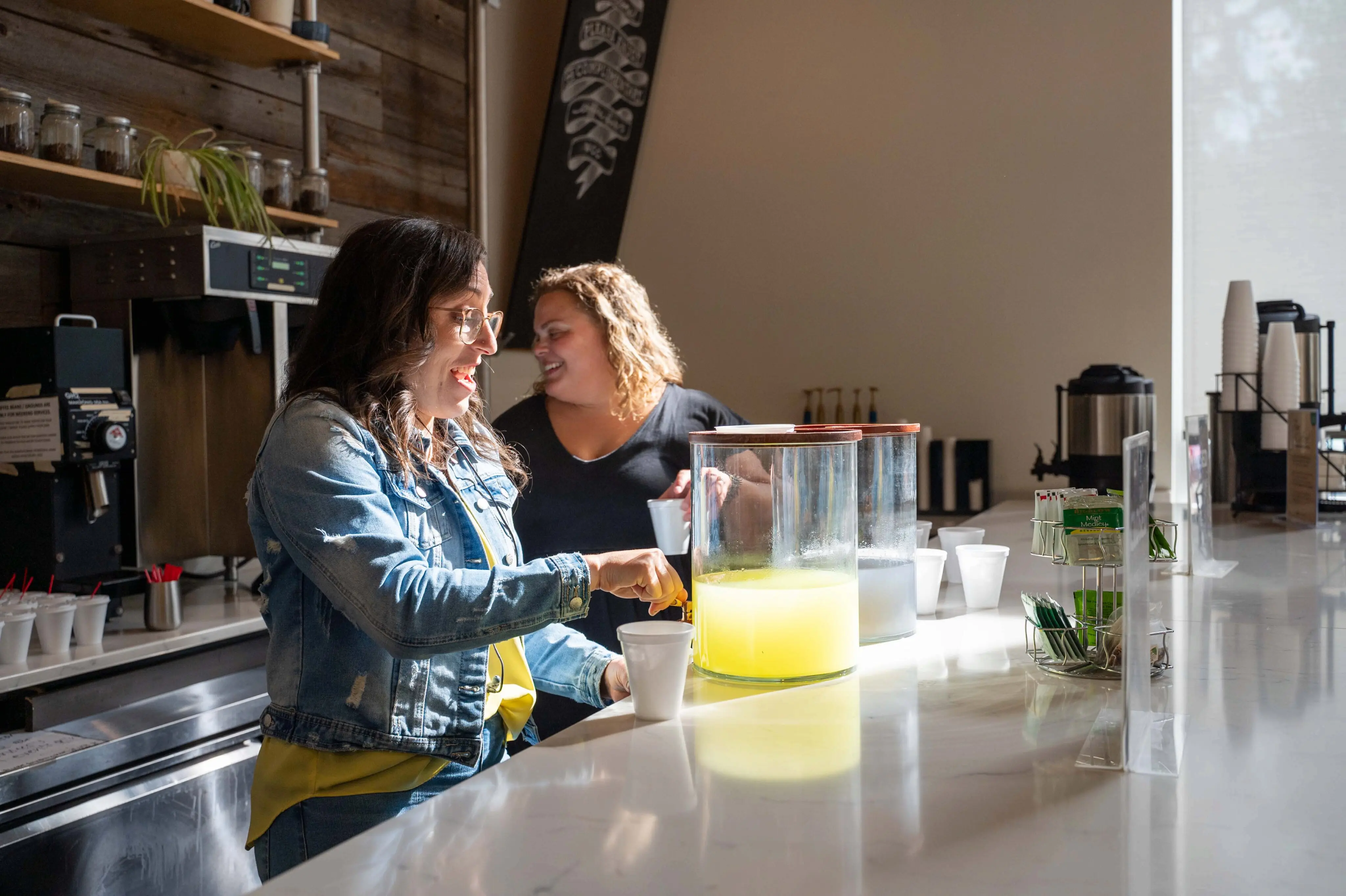 Two women smile and chat while standing behind a white marble counter at the Walnut Creek Downtown cafe area. One woman, wearing a denim jacket, is pouring a bright yellow beverage from a large glass dispenser into a white cup. The modern space features rustic wood-paneled walls, industrial shelving with coffee jars, and coffee-making equipment in the background. Natural sunlight streams in from a large window on the right, illuminating the scene.