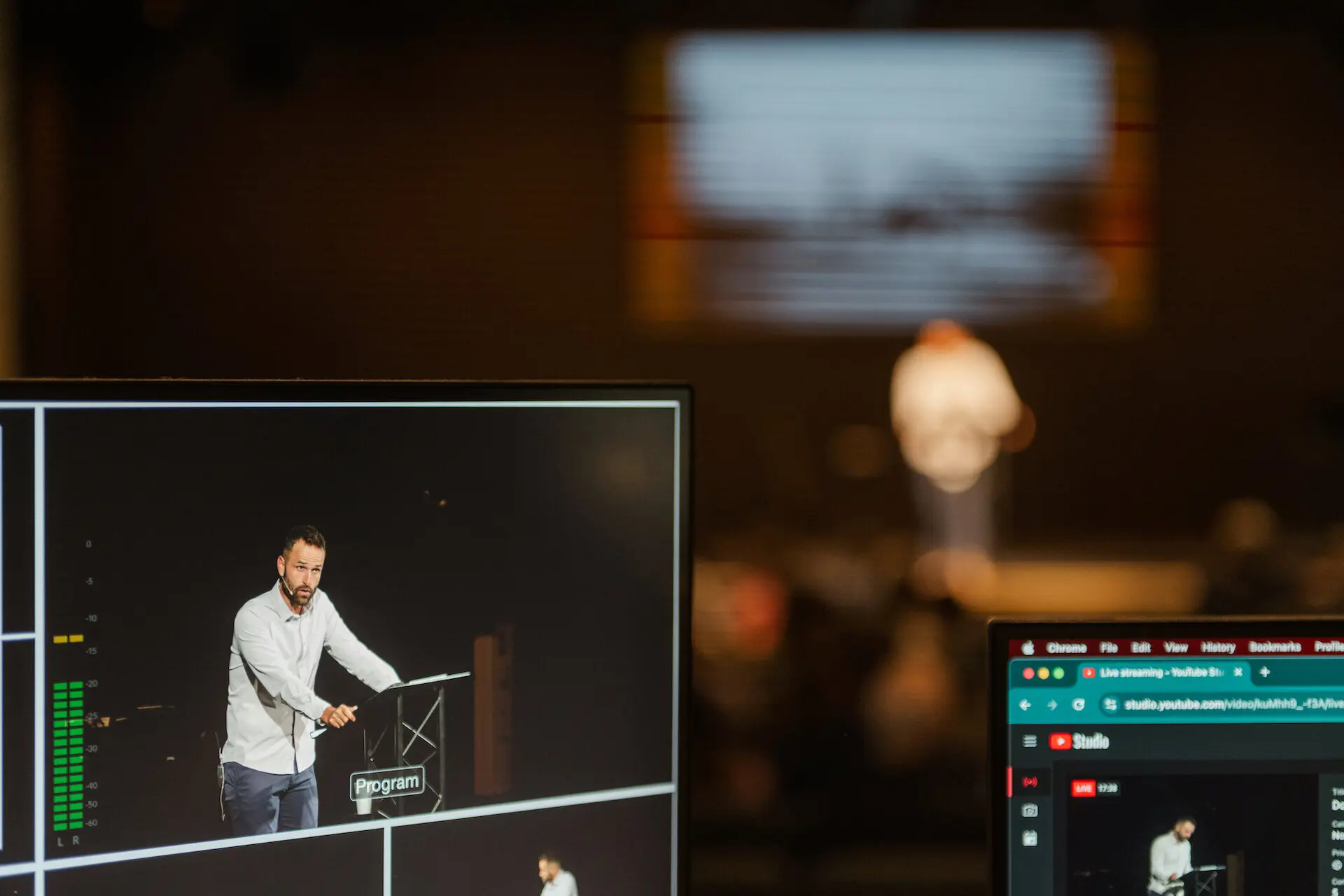 A behind-the-scenes look at the live stream production for a service at Walnut Creek Downtown. In the foreground, a large production monitor shows a clear, live feed of a man speaking at a podium. To the right, a smaller laptop screen displays the YouTube Studio interface, showing the "Live" broadcast status. In the soft-focus background, the actual speaker can be seen on stage in the dimly lit auditorium, creating a depth-of-field effect that highlights the technology used to broadcast the service.