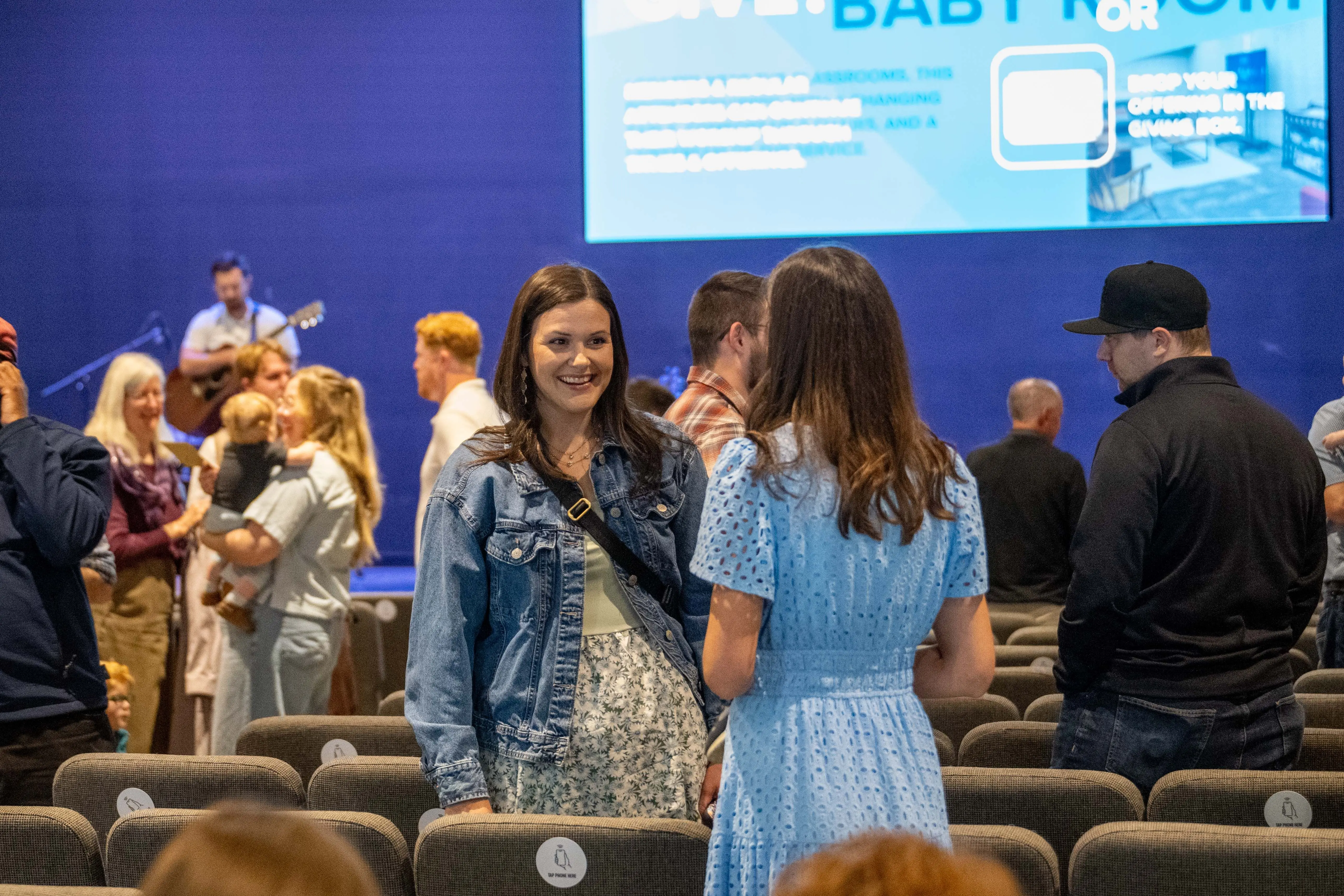 A candid photo of two women talking and smiling in the auditorium of Walnut Creek Downtown. One woman wears a denim jacket over a floral dress, while the other is in a light blue eyelet dress. They are standing among rows of grey cushioned chairs. In the background, other church members are socializing, including a woman holding a young child. A large blue screen displays information about "Baby Room" and "Giving," and a musician with a guitar is partially visible on the stage.