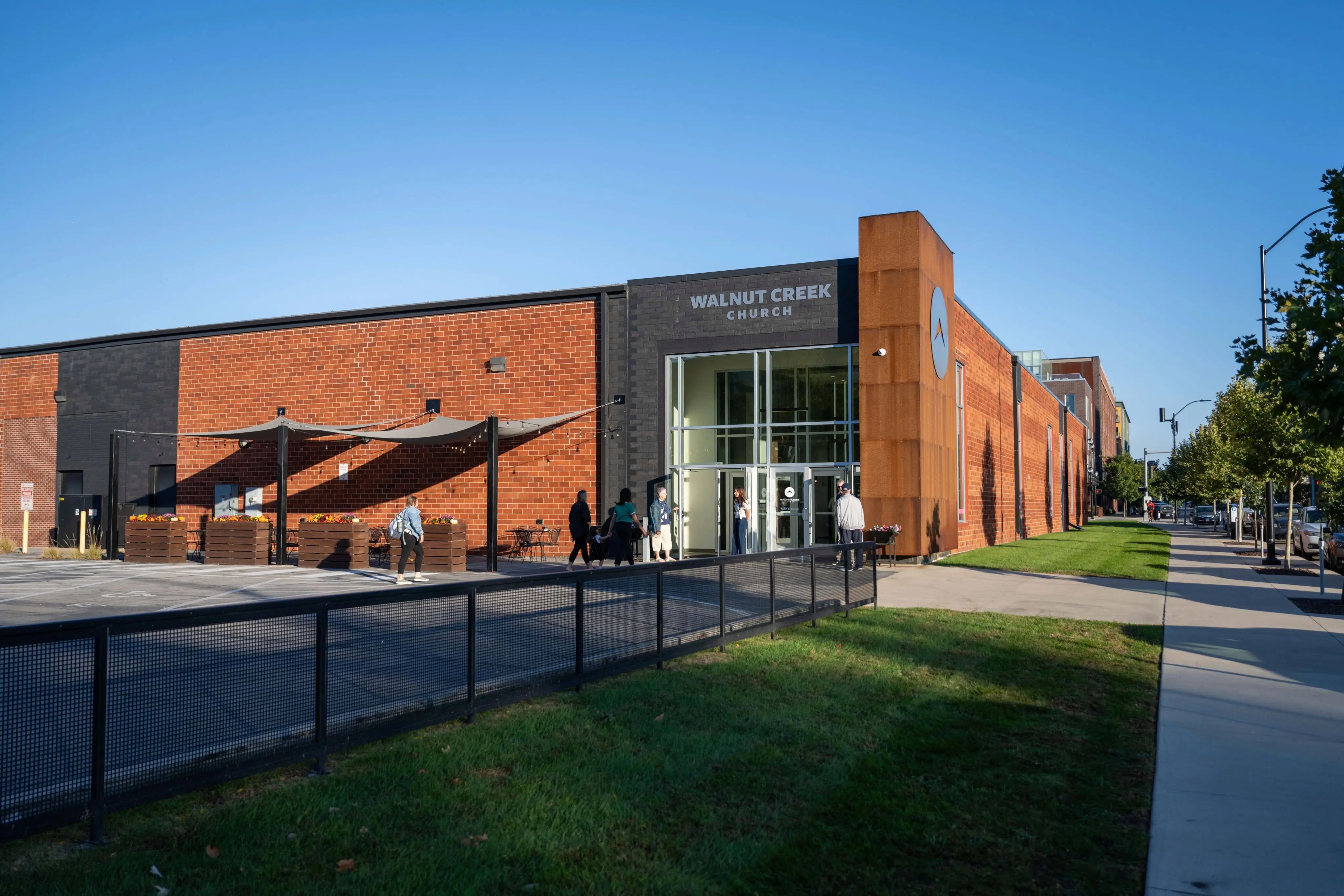 The exterior of Walnut Creek Church on a clear, sunny day. The building features a modern industrial design with red brick walls, black accents, and a prominent rusted-metal tower bearing the church’s circular logo. A large glass entryway serves as the main focal point, where several people are seen entering. To the left, an outdoor patio area is covered by tan sun sails and decorated with wooden planter boxes. A clean concrete sidewalk and a small green lawn line the front of the building under a bright blue sky.
