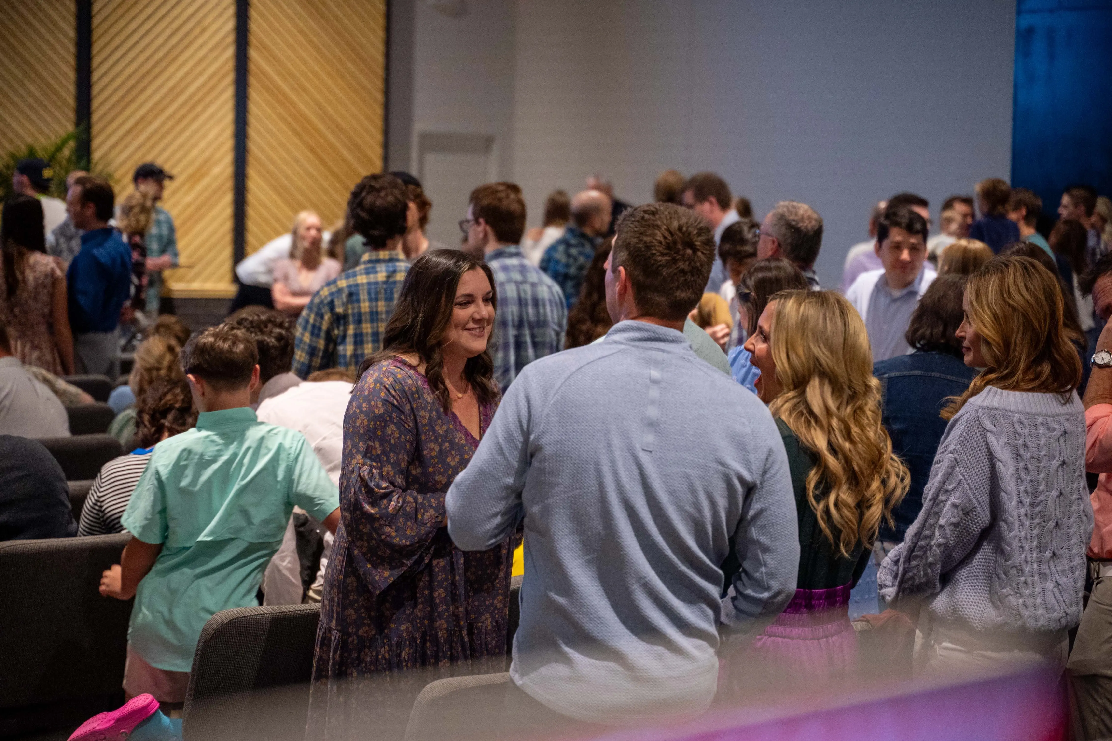 A lively crowd of people socializes in the auditorium of Walnut Creek Downtown. In the center, a woman in a purple floral dress smiles warmly while talking to a man in a light blue quarter-zip pullover. Around them, other church members of various ages are gathered in groups, chatting between rows of grey upholstered chairs. The background features a large wall with a decorative light-wood chevron pattern, and the overall atmosphere is bright and communal.