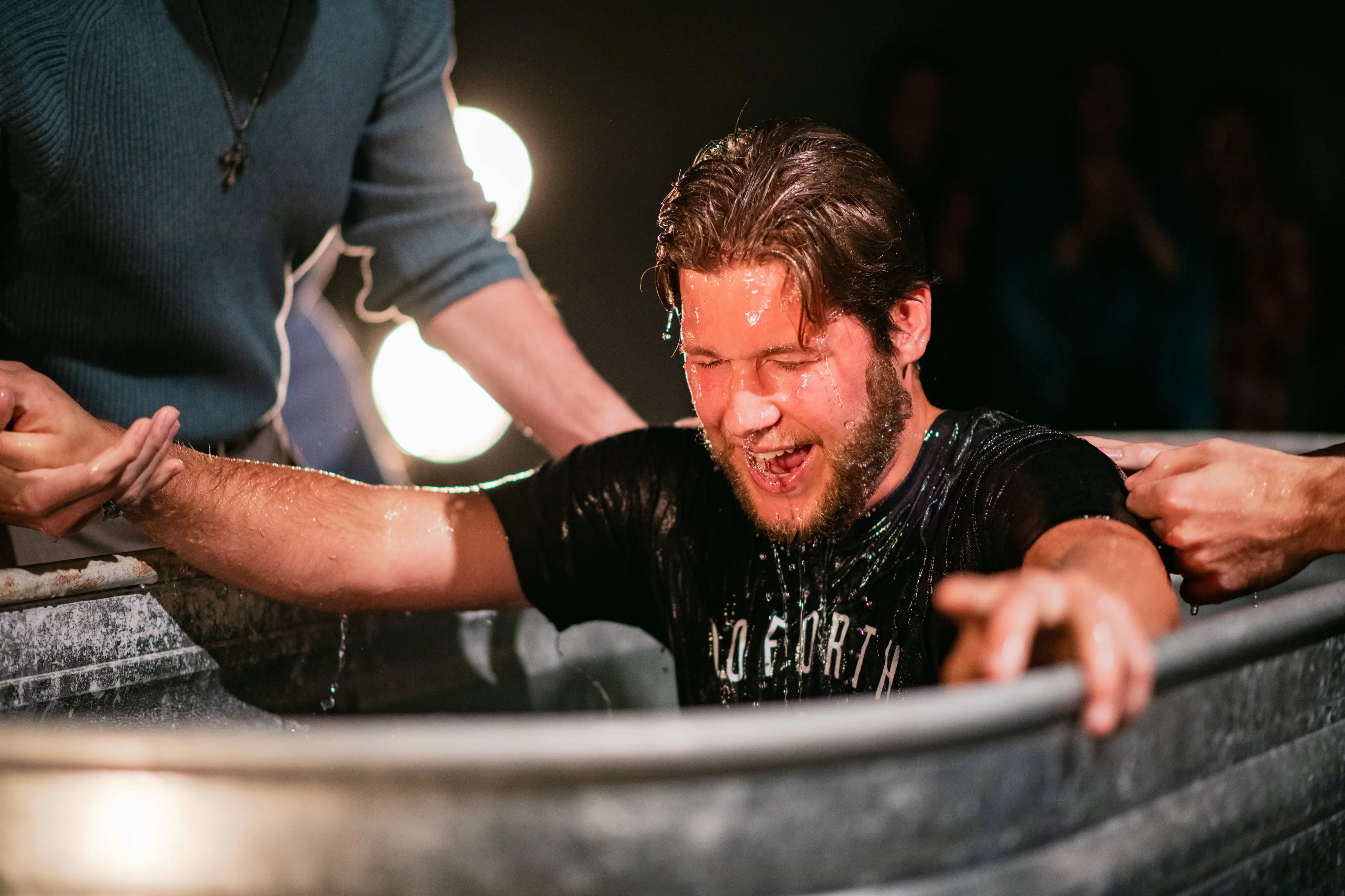 A man with a beard and wet hair emerges from a metal trough during a baptism at Walnut Creek Downtown. He has an expression of pure joy and excitement as water drips from his face and black t-shirt. Other members of the church reach out to support his arms and shoulders, with soft, glowing stage lights creating a warm atmosphere in the background.