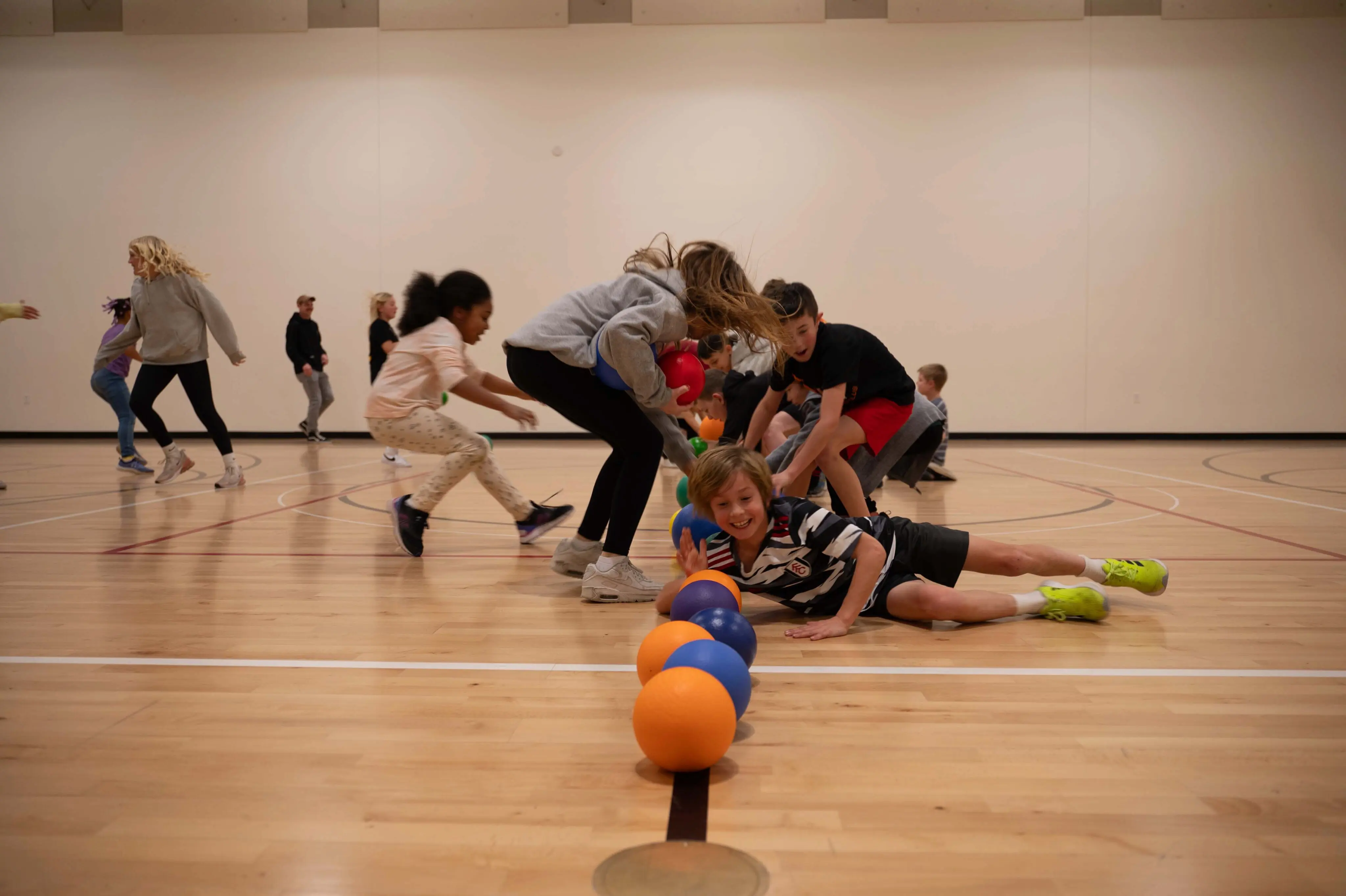 A high-energy game of dodgeball taking place in a gymnasium at Walnut Creek Downtown. A line of colorful rubber balls—orange, blue, and purple—sits along the center court line. In the center, a young boy in a striped jersey dives toward the balls with a big smile, while other children and teenagers rush forward from both sides to grab them. The background shows more participants in motion against white gym walls, capturing the excitement of a youth group activity.