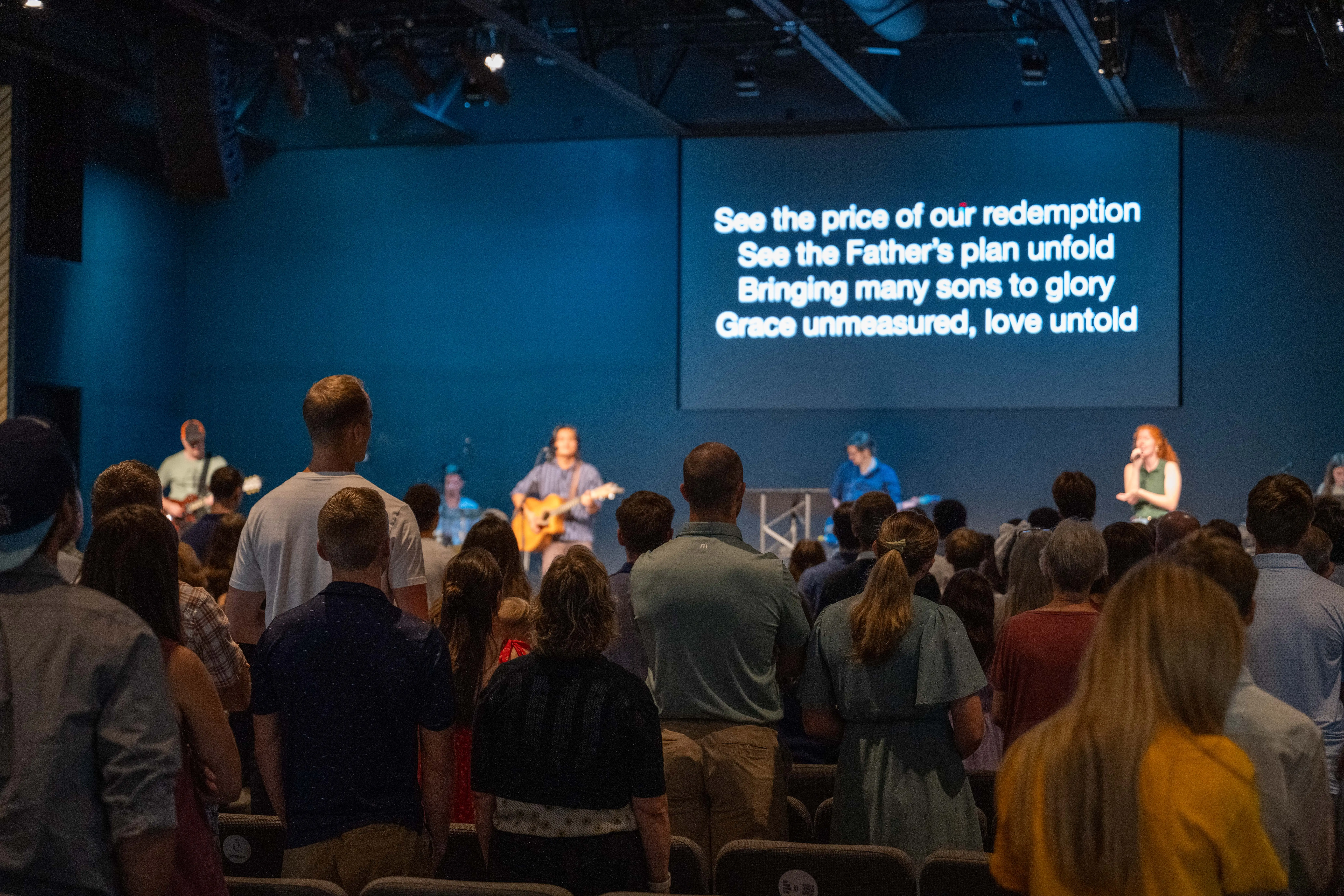 A wide-angle view of a Sunday service at Walnut Creek Downtown. A speaker stands at a podium on a raised, curved stage, addressing a large, attentive congregation seated in rows of grey chairs. To the right of the speaker, a drum set and musical equipment are visible on stage, and a large screen overhead displays the Bible verse 1 Peter 1:23-25. The auditorium has a modern, industrial feel with an exposed black ceiling, recessed lighting, and dark brick accents along the stage.