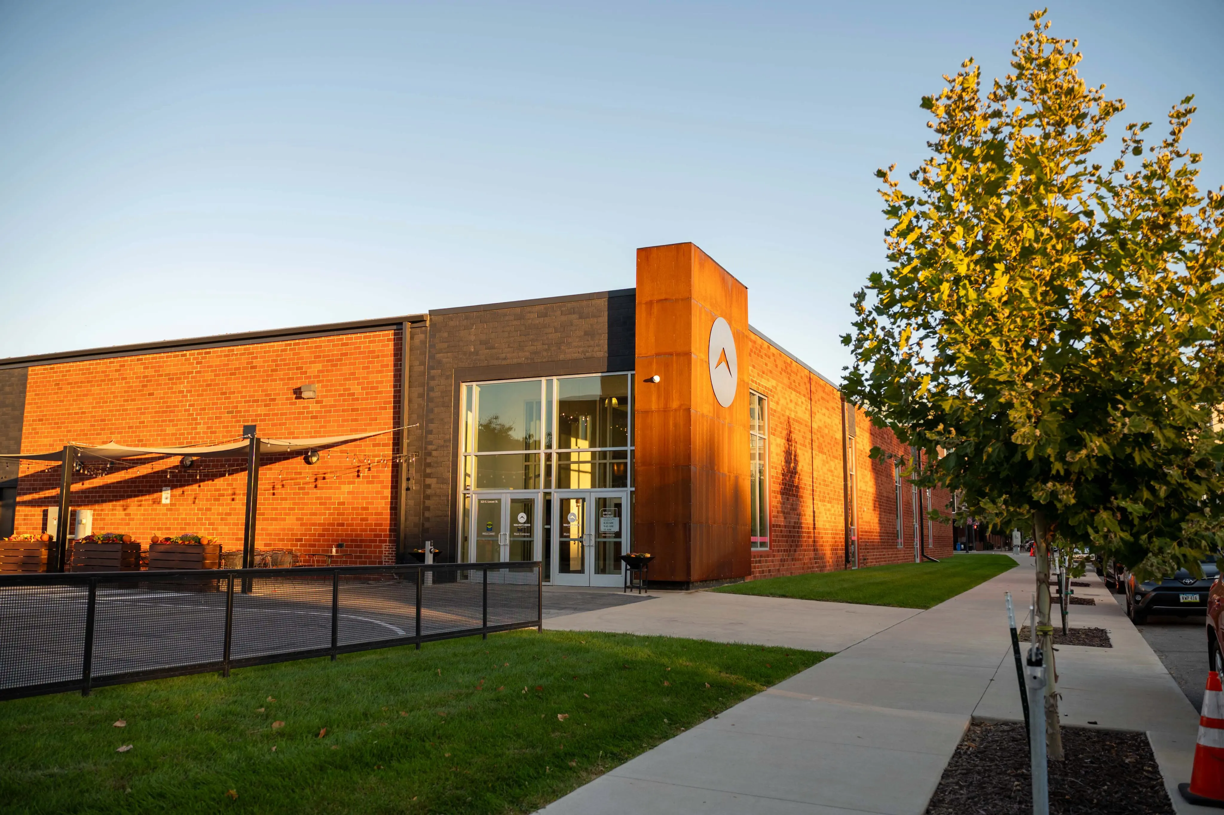 The exterior of the Walnut Creek Downtown church building during a golden hour sunset. The modern architecture features a combination of red brick, dark stone, and a prominent rusted metal tower displaying the church’s circular logo. Large glass doors and windows mark the entrance, and an outdoor patio area with string lights is visible to the left. A sidewalk lined with young trees leads past the building under a clear blue sky.