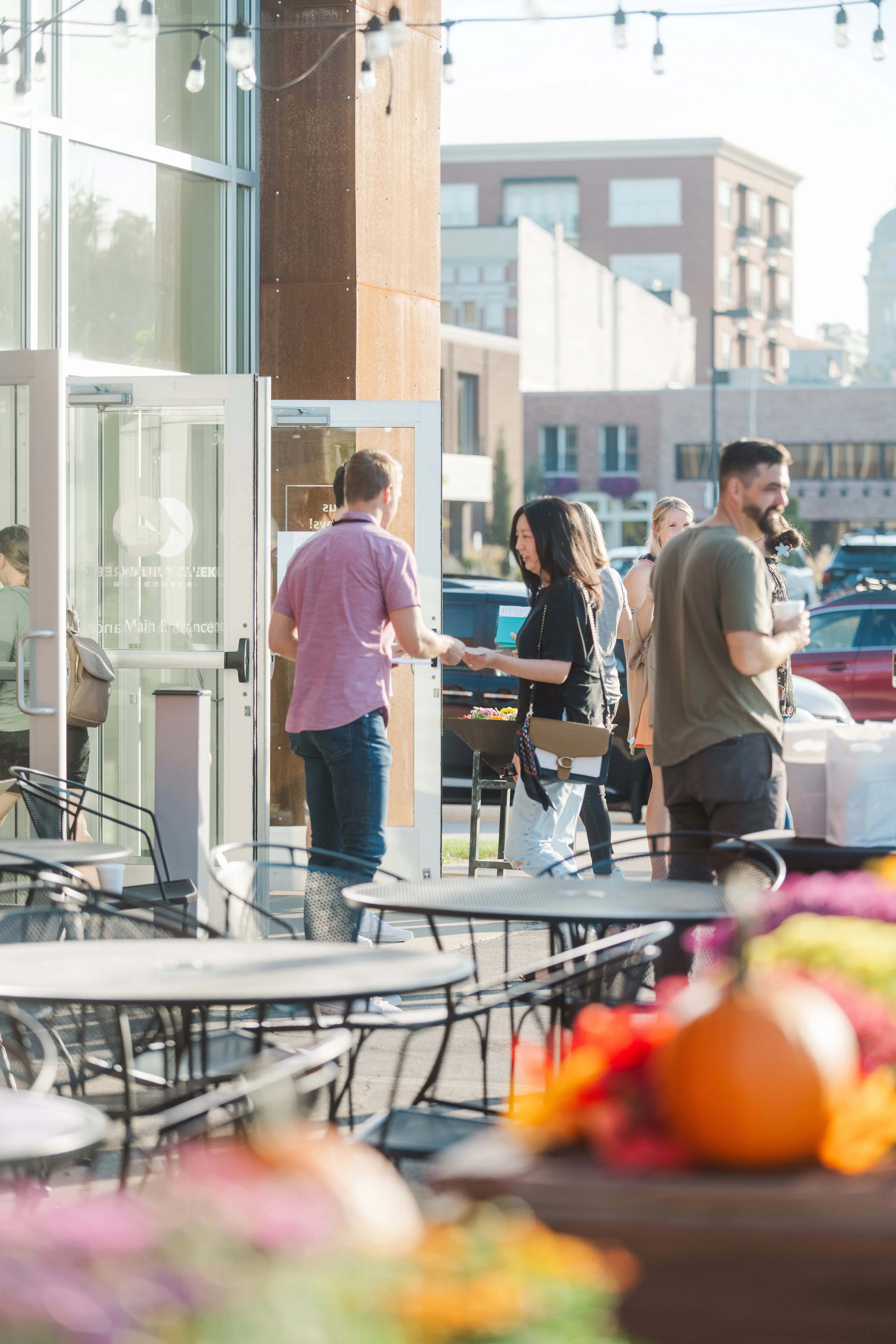 A welcoming scene at the main entrance of Walnut Creek Church Downtown, featuring diverse community members gathering outdoors. A greeter in a pink shirt welcomes visitors near the glass doors, while others enjoy coffee at patio tables decorated with flowers in a sunny, urban setting.