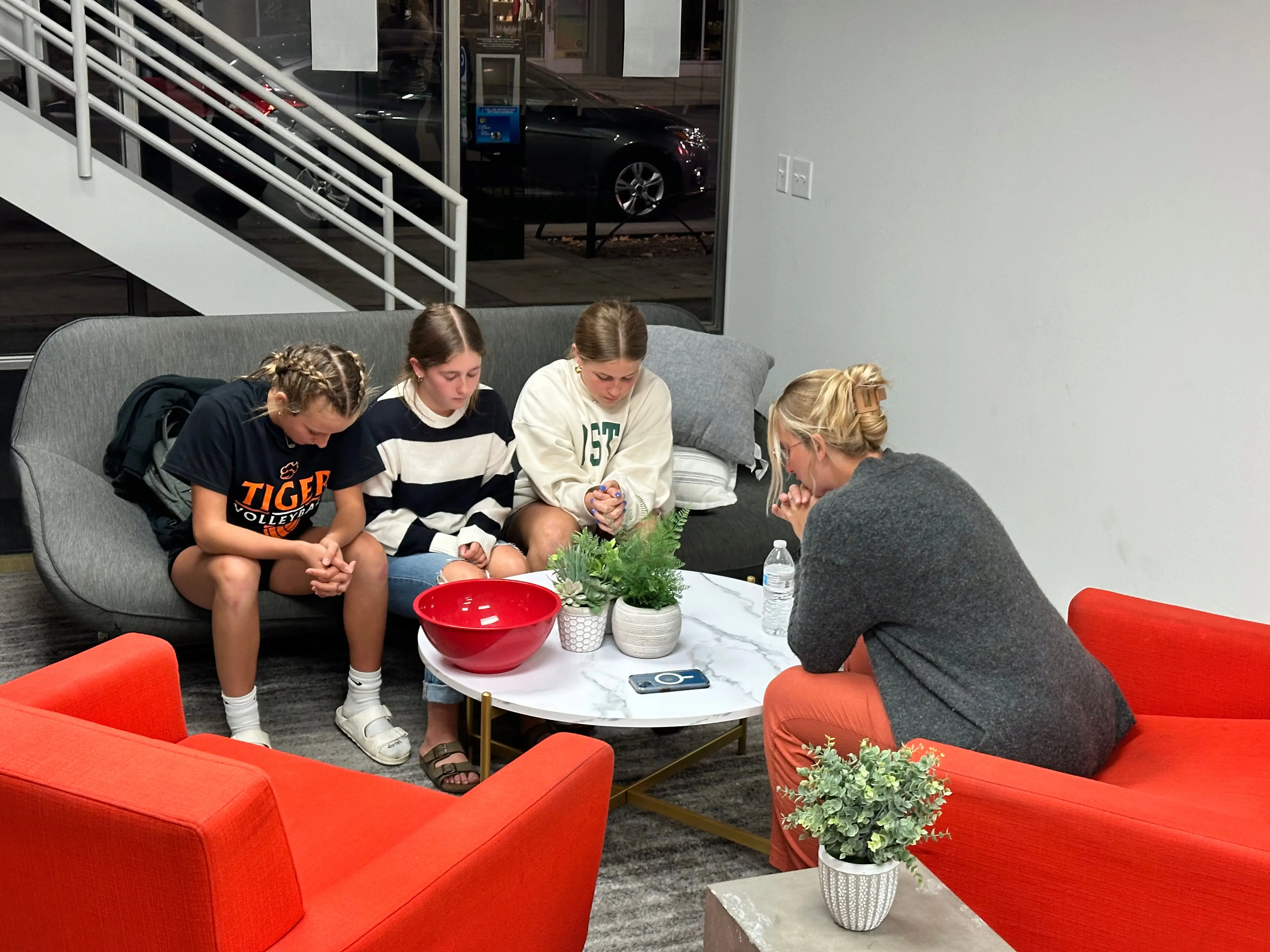 A small group of four young women gathered in prayer at Walnut Creek Downtown. Three girls sit on a modern grey sofa, while a fourth sits opposite them in an orange armchair. They are all leaning forward with their heads bowed and hands clasped. Between them is a white marble coffee table holding a large red bowl and small potted plants. The setting is a clean, contemporary lobby area with a white staircase and a large window in the background.