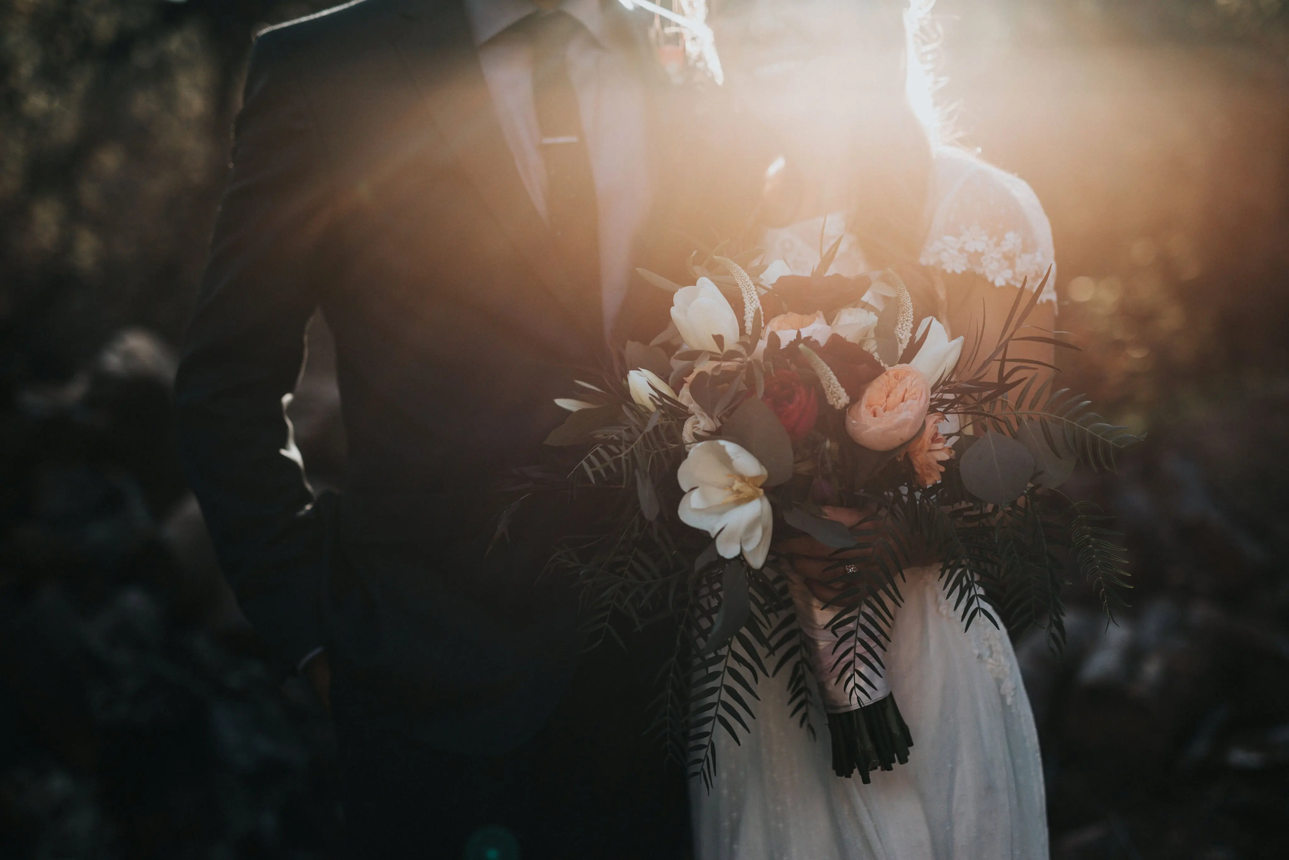 A close-up, moody photograph of a bride and groom standing together at sunset. The bride holds a lush bouquet featuring white tulips, peach roses, and dark red flowers mixed with various textured greenery. Golden sunlight creates a bright lens flare that partially obscures their faces, highlighting the bride's white lace gown and the groom's dark suit and tie. The background is softly blurred with dark, natural tones.