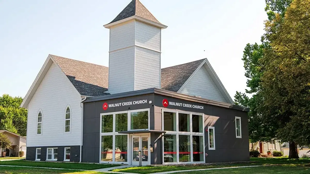 A photograph of the Walnut Creek Church in Altoona. The building combines a traditional white chapel with a steeple and a modern, dark grey addition featuring large glass windows and the church logo. The text below lists the address as 107 6th St SW, Altoona, IA, with service times on Sundays at 9:30 a.m.