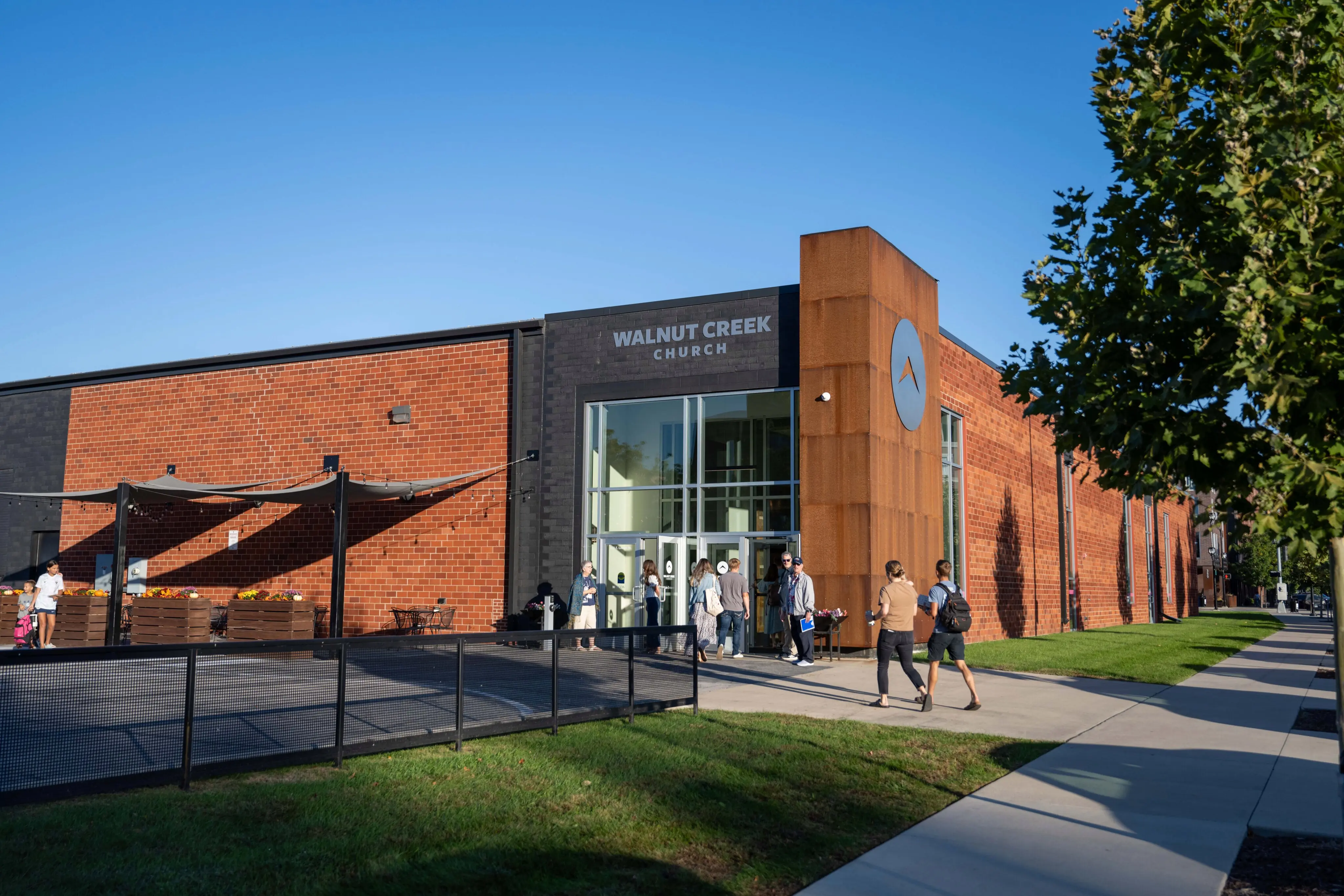 A photograph of the Downtown Walnut Creek Church, a large industrial-style brick and glass building with a prominent rusted-metal architectural feature holding the church logo. People are seen walking toward the modern glass entrance. The text below lists the address as 323 E. Locust St. Des Moines, IA, with Sunday service times at 8:30 a.m., 9:45 a.m., and 11:00 a.m.