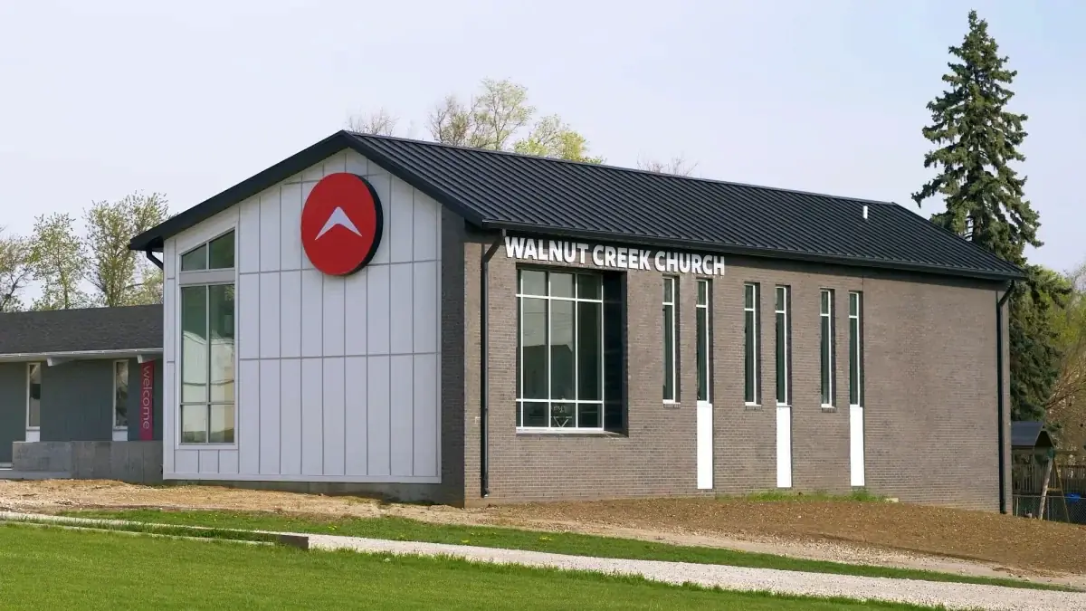 A photograph of the South location of Walnut Creek Church. The building is a modern, single-story structure with grey brick, vertical white paneling, and a large red circular logo. It features several tall, narrow windows and a black metal roof.