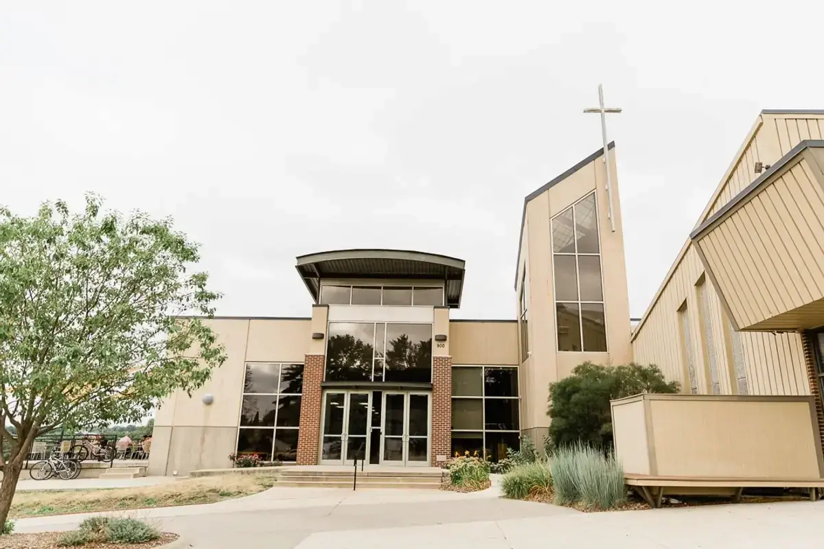 A photograph of the Windsor Heights location of Walnut Creek Church. The facility is a large, tan-colored multi-wing building with a modern entrance under a curved overhang and a tall, thin cross visible on the roofline against a bright sky.
